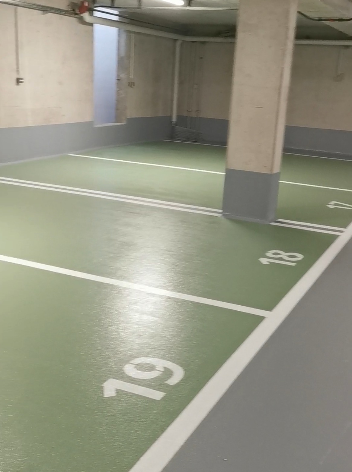 Oznakowane, zielone stanowiska parkingowe w garażu podziemnym z szarą posadzką, wykończone posadzki z żywicy epoksydowej. Numeracja stanowisk widoczna na podłodze.