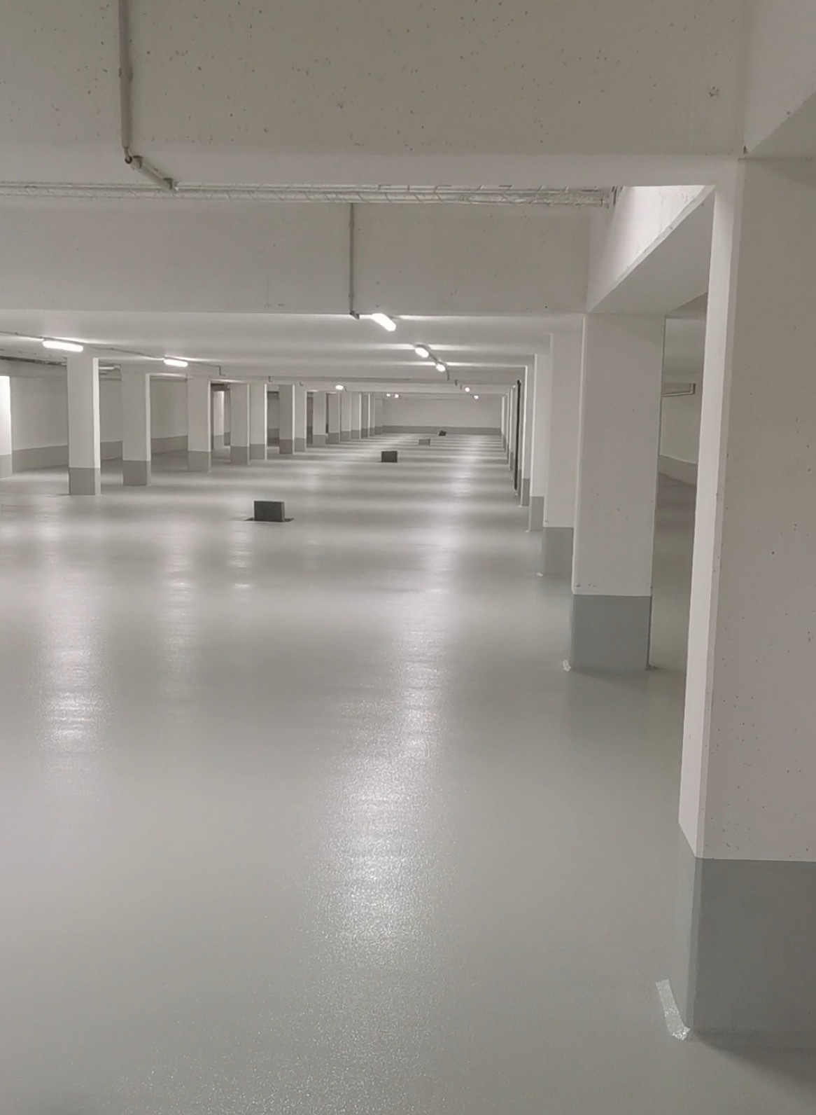 Jasnoszara, błyszcząca posadzka w hali z rzędami kolumn. Widoczne lampy sufitowe i fragmenty taśmy zabezpieczającej przy kolumnach. Perspektywa zbieżna w głąb.