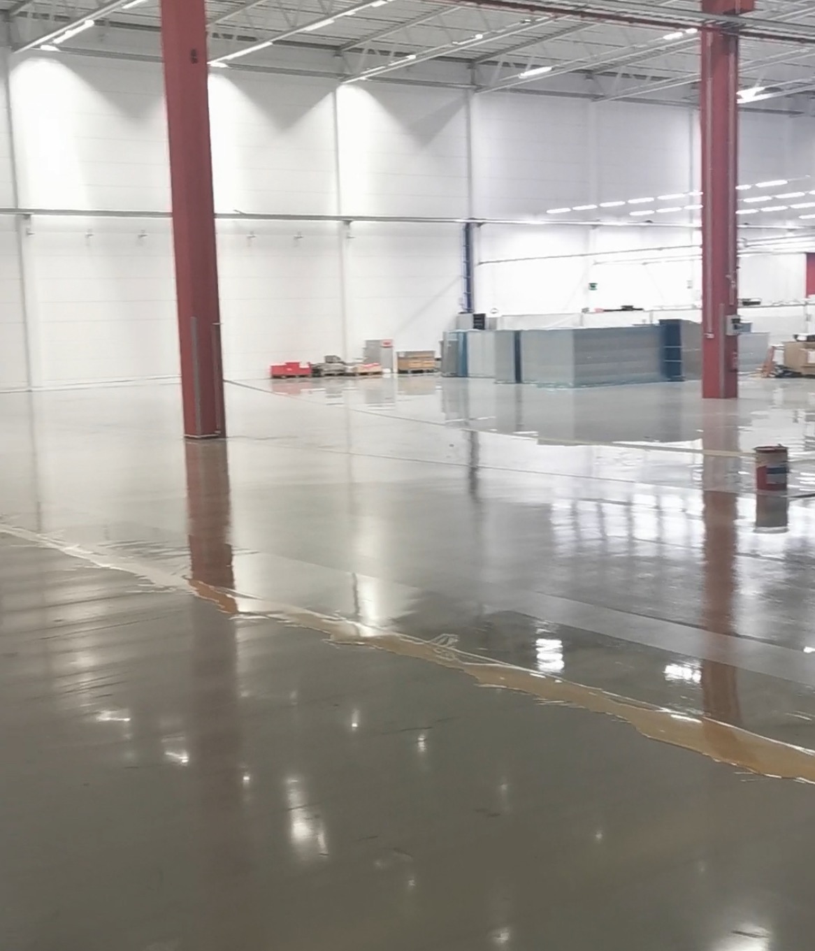 Błyszcząca, szara posadzka w hali przemysłowej z widocznymi czerwonymi słupami i materiałami budowlanymi w tle. Świeżo wylana, mokra powierzchnia.