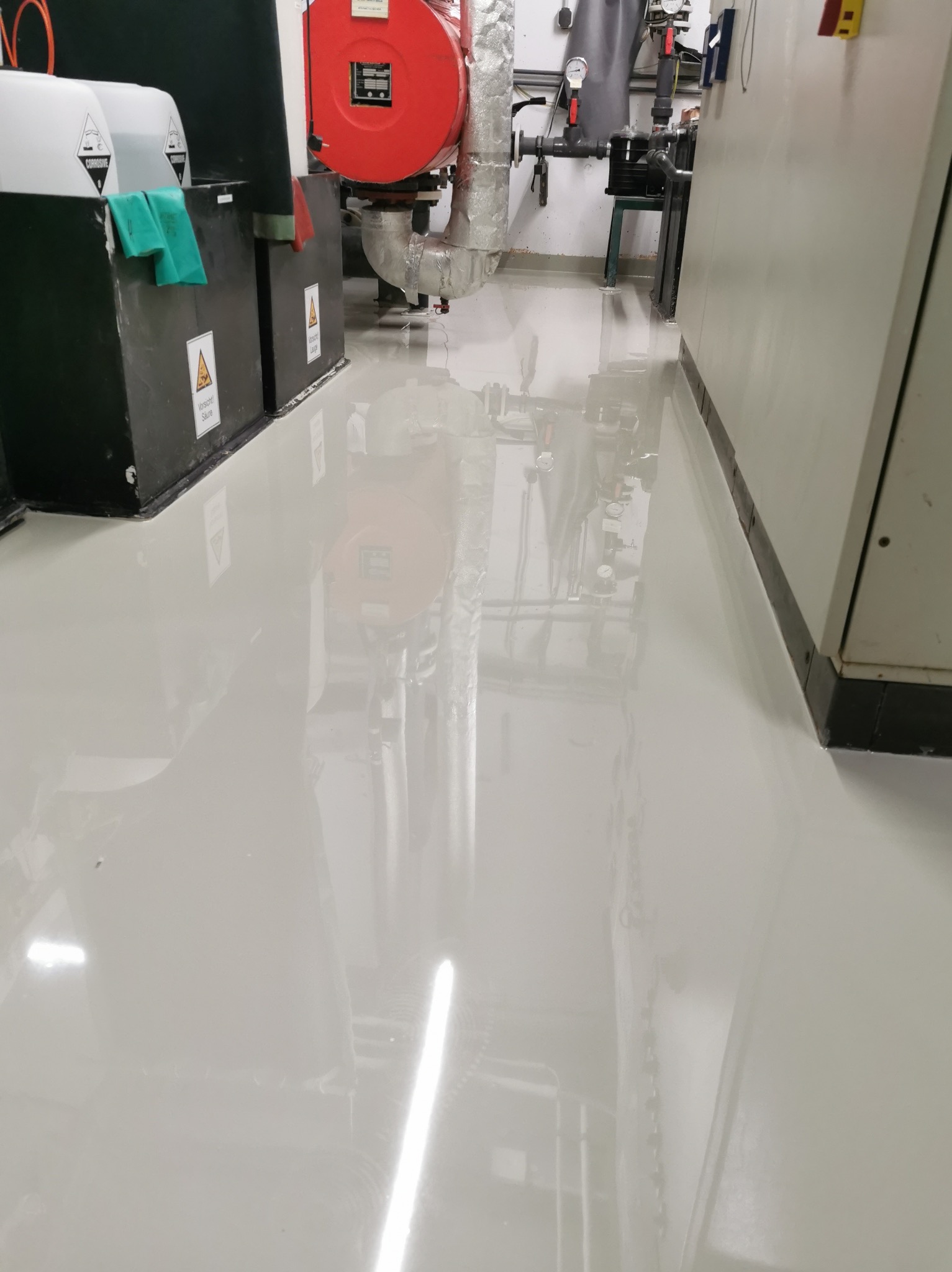 Błyszcząca, jasnoszara posadzka żywiczna w pomieszczeniu technicznym z instalacjami. Realizacja w Resko. Widoczne pojemniki z oznaczeniami ostrzegawczymi.