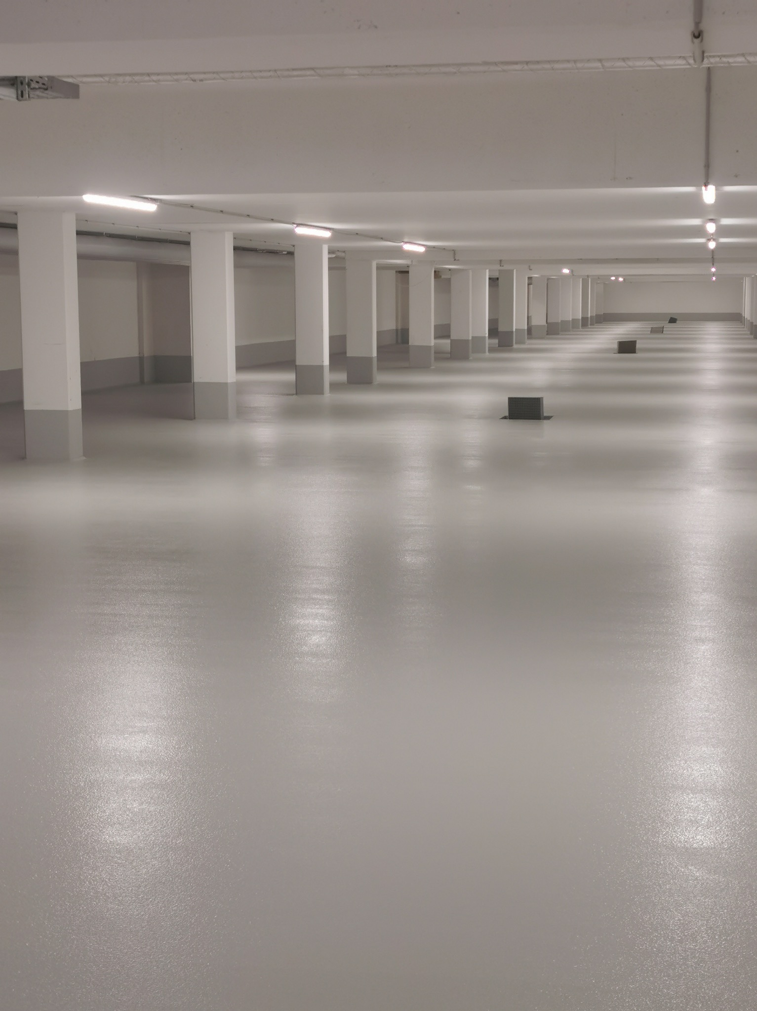 Jasnoszara, gładka posadzka w hali z rzędami kolumn i oświetleniem. Widoczne kratki wentylacyjne w podłodze. Perspektywa zbieżna, podkreślająca przestrzeń.