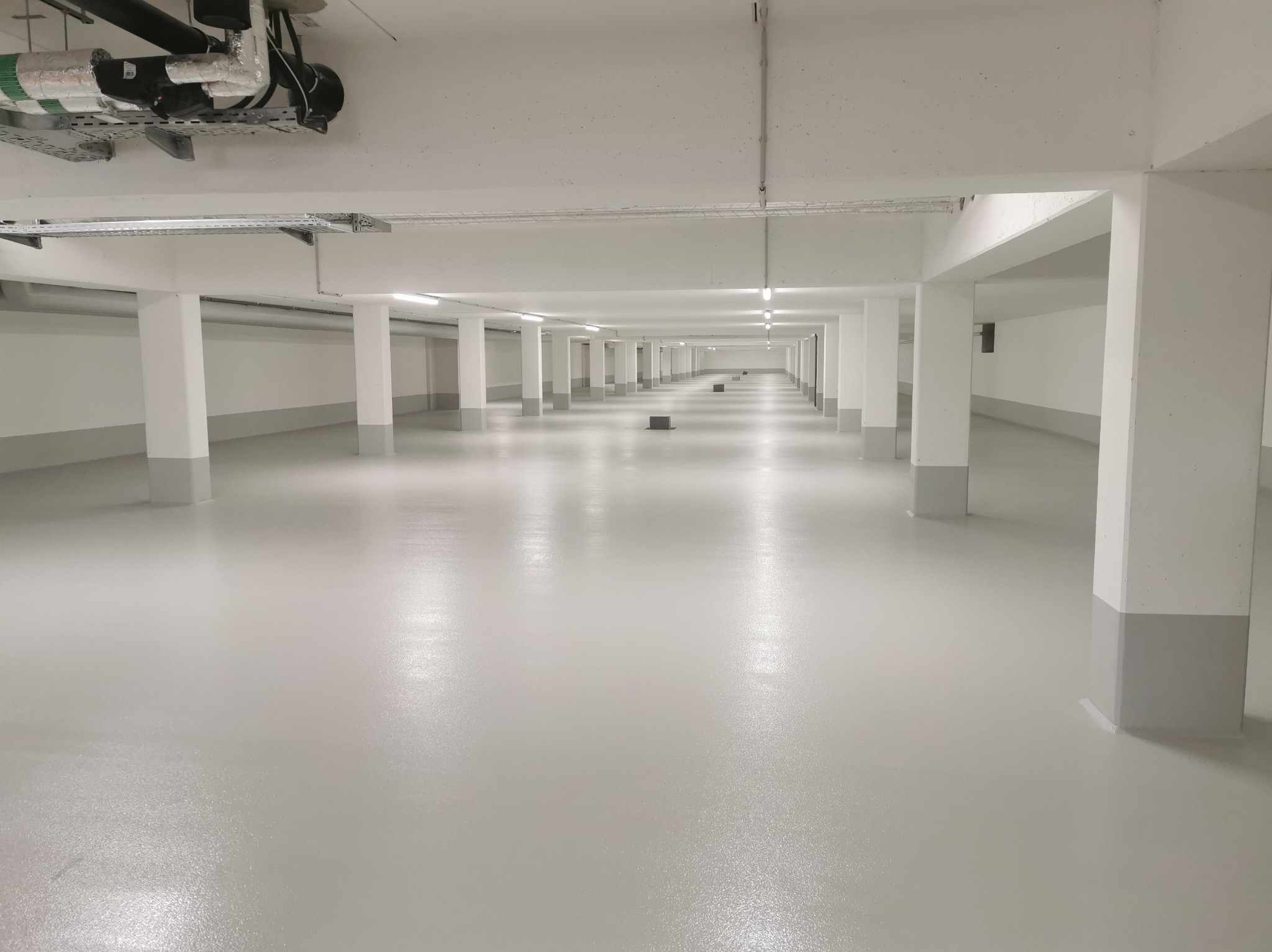 Jasnoszara, gładka posadzka w hali magazynowej z rzędami kolumn. Widoczne instalacje pod sufitem. Perspektywa zbieżna, podkreślająca przestrzeń.