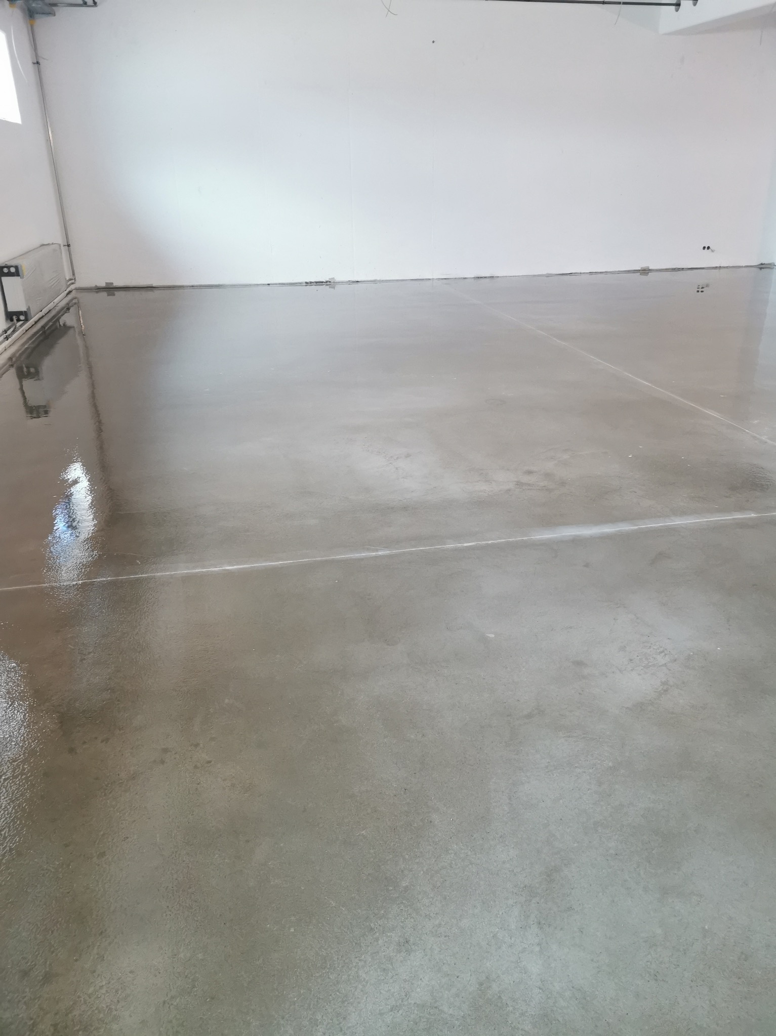 Błyszcząca, szara posadzka w jasnym pomieszczeniu. Widoczne delikatne refleksy światła i linie podziału. Minimalistyczne wnętrze z gładką powierzchnią podłogi.