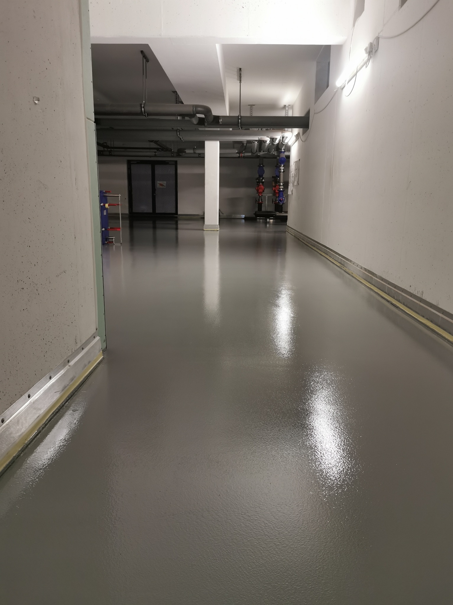 Gładka, szara posadzka w hali przemysłowej z widocznymi instalacjami pod sufitem i metalowymi listwami przypodłogowymi. Połyskująca powierzchnia odbija światło.