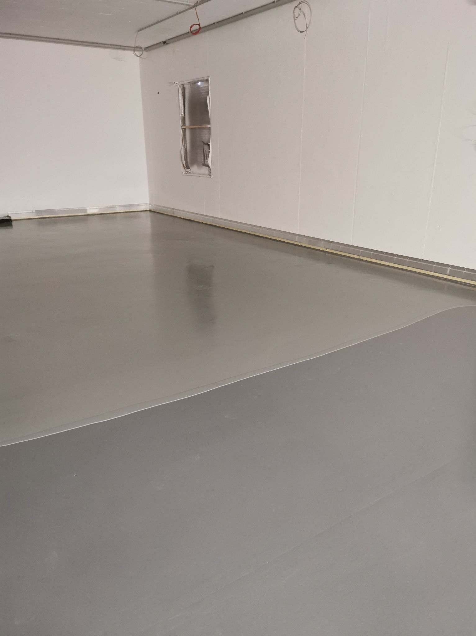 Szara, gładka posadzka w jasnym pomieszczeniu przemysłowym. Widoczne łączenia paneli ściennych i zabezpieczone okno. Minimalistyczny, surowy styl.