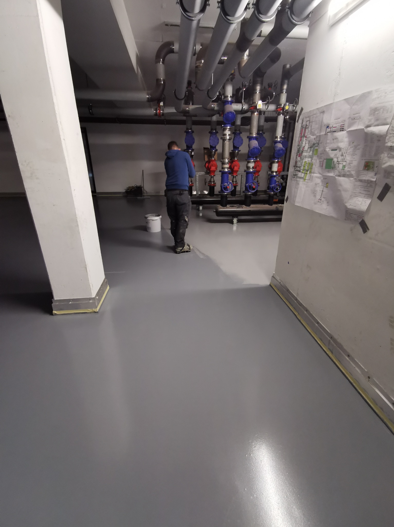 Aplikacja szarej, błyszczącej posadzki w pomieszczeniu technicznym z rurami. Pracownik w niebieskiej bluzie maluje podłoże. Na ścianie widoczny schemat instalacji.
