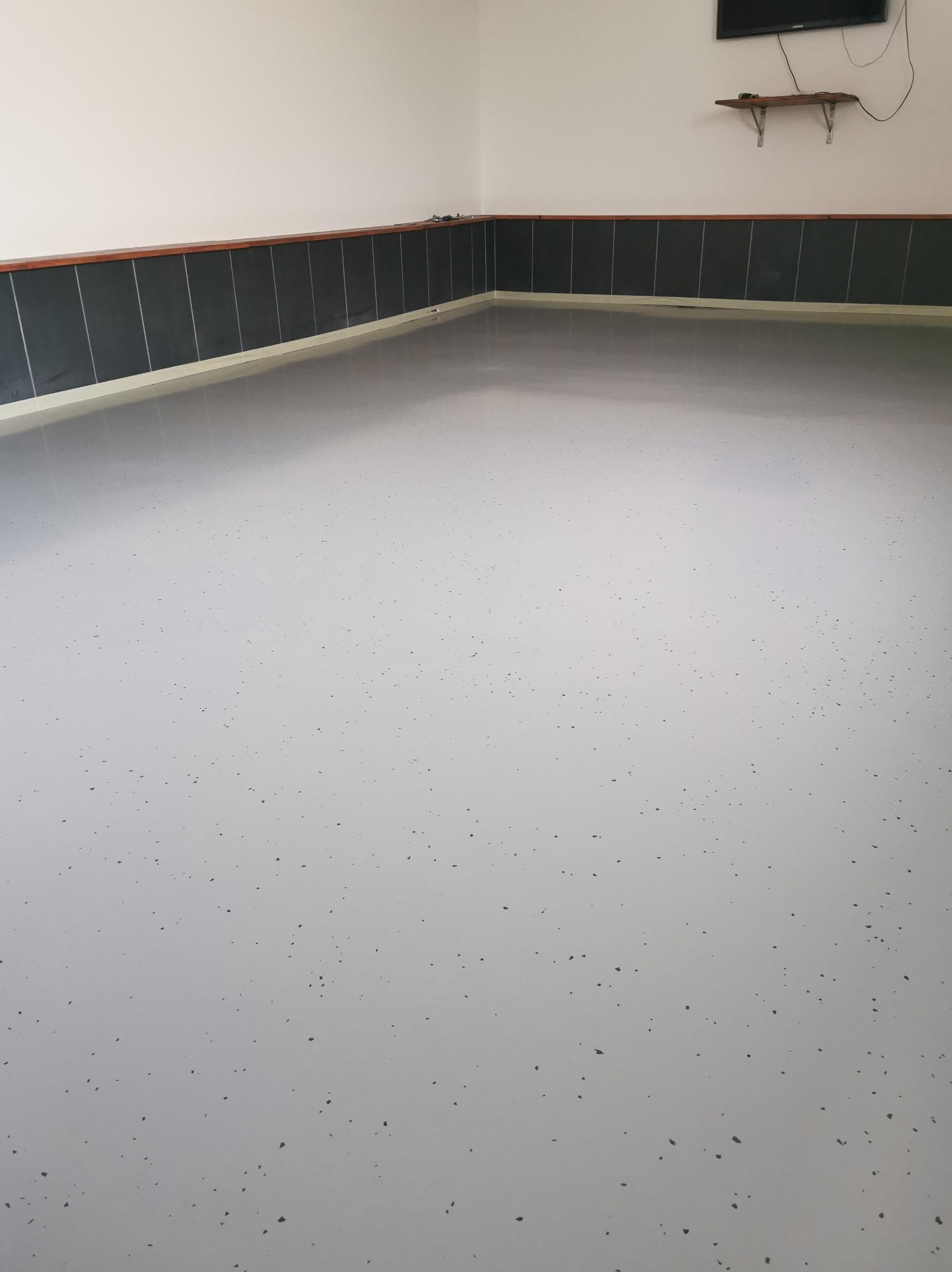 Jasnoszara posadzka żywiczna z ciemnymi drobinkami w garażu, widoczna dolna część ściany z ciemnymi panelami i białą listwą przypodłogową. Na ścianie półka i telewizor.