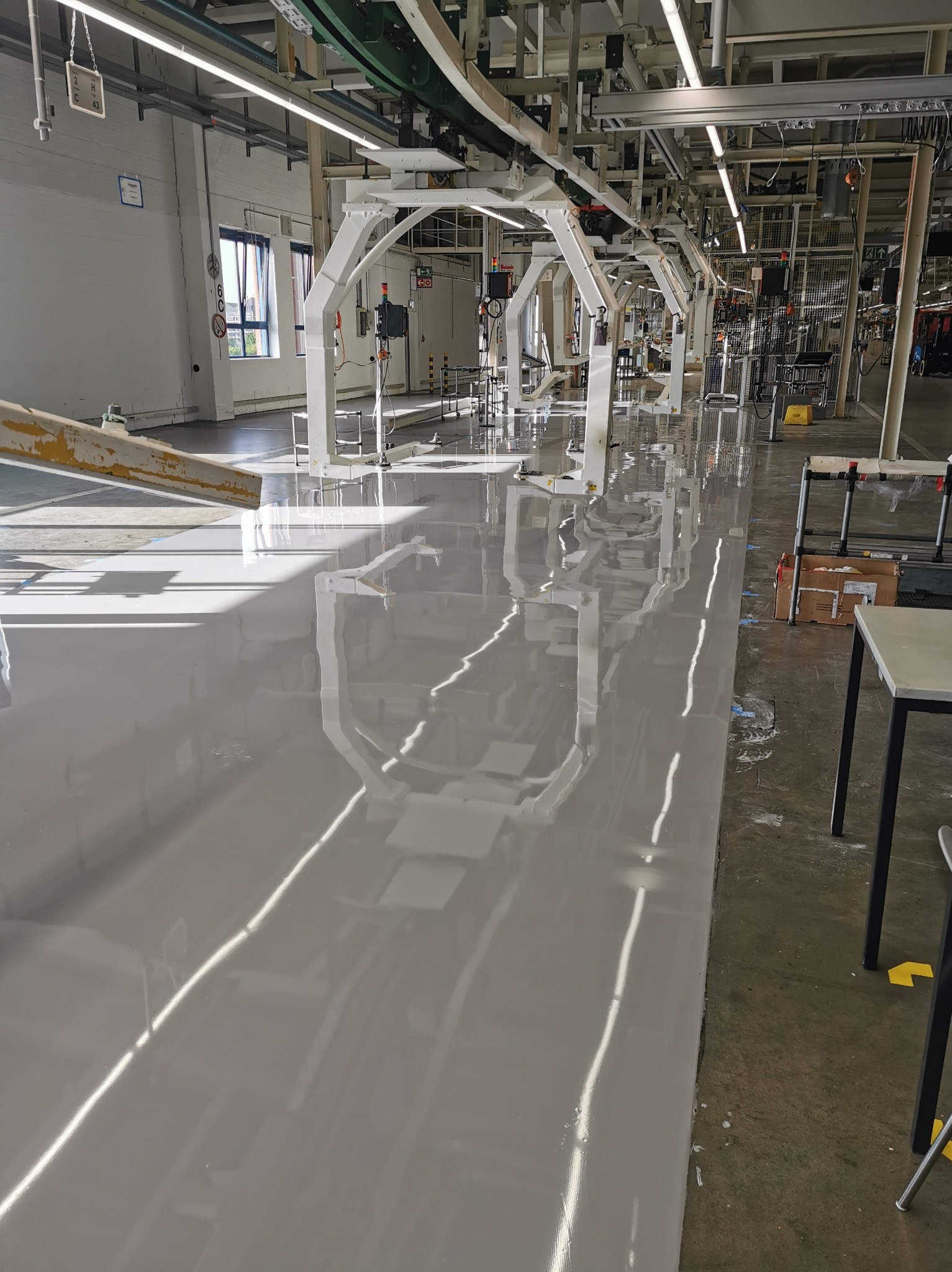 Błyszcząca, szara posadzka w hali produkcyjnej z białymi konstrukcjami sufitowymi i oknami w tle. Widoczne refleksy świetlne na gładkiej powierzchni.