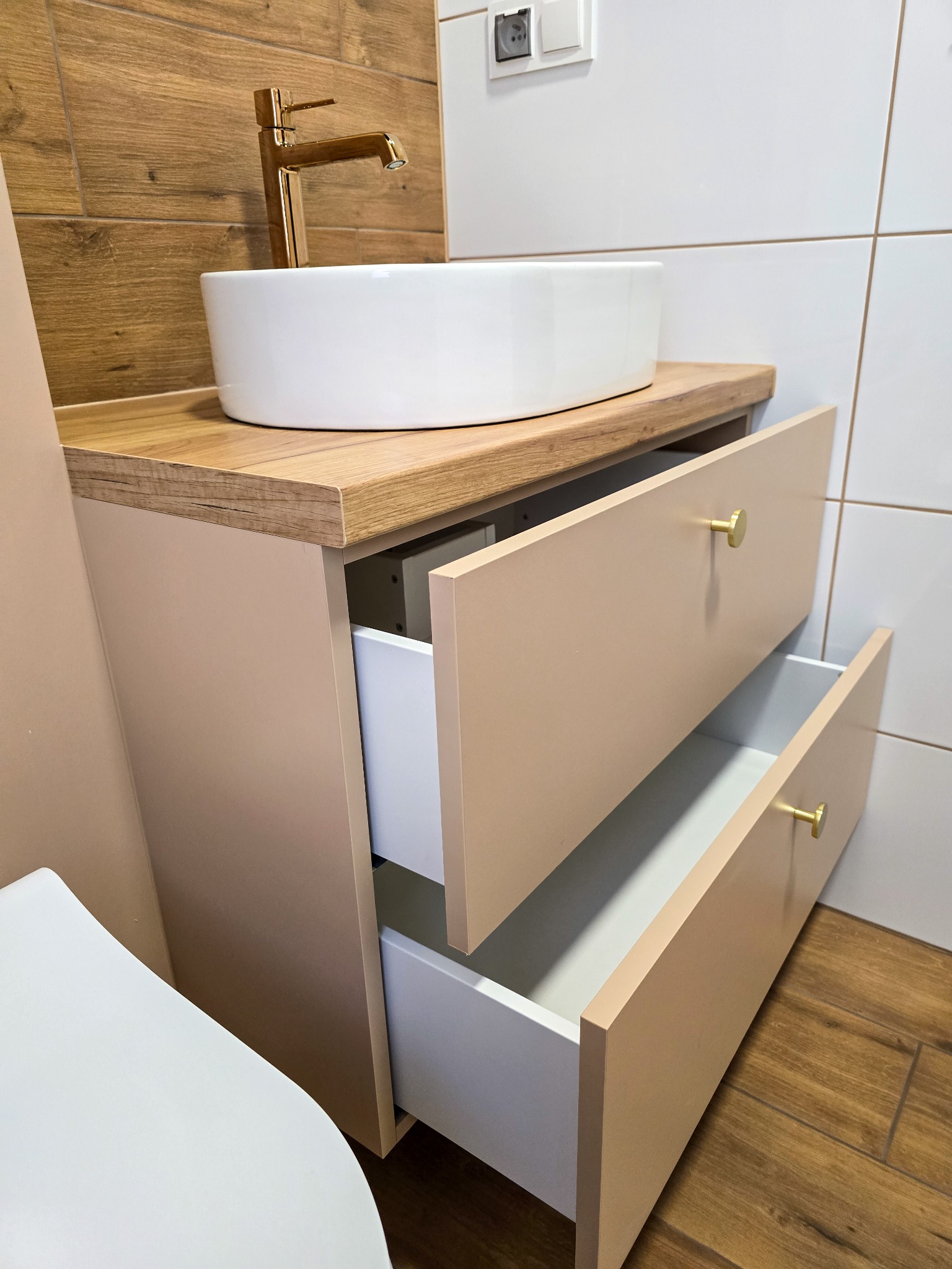 Nowoczesna szafka łazienkowa z otwartymi szufladami, drewnianym blatem i białą umywalką nablatową, złota bateria, białe i drewniane płytki w tle.