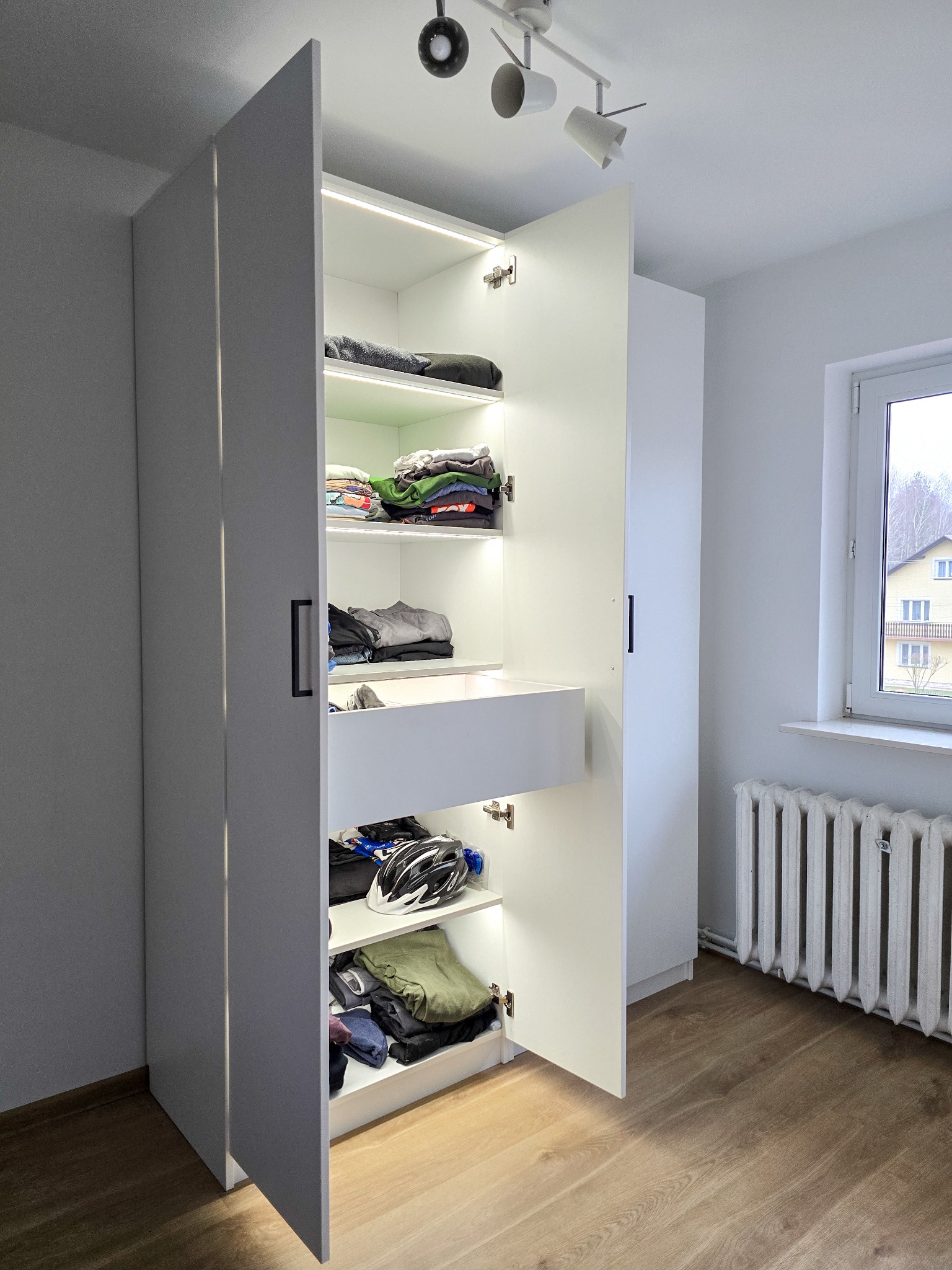 Biała szafa z oświetleniem LED, częściowo otwarta, ukazująca ubrania i szufladę. Widoczne czarne uchwyty, parkiet, okno i grzejnik w tle.