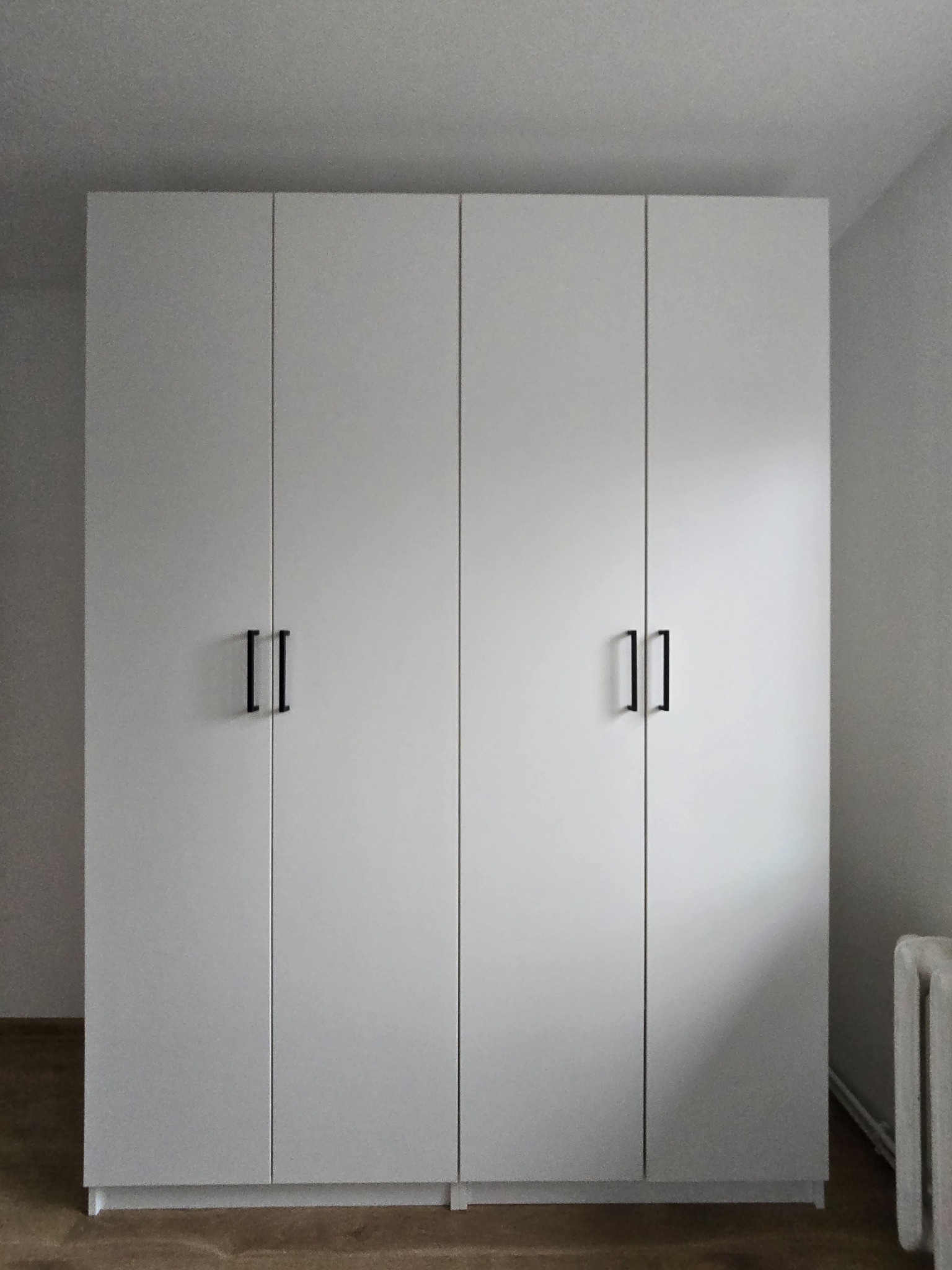 Biała szafa czterodrzwiowa z czarnymi, minimalistycznymi uchwytami, ustawiona na drewnianej podłodze w jasnym pomieszczeniu. Widoczny fragment grzejnika.