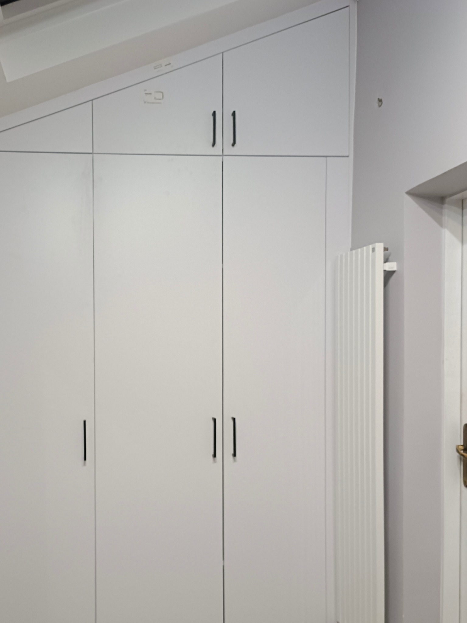 Biała szafa wnękowa z czarnymi uchwytami, dopasowana do skośnego sufitu, obok grzejnik i fragment drzwi. Minimalistyczny design.