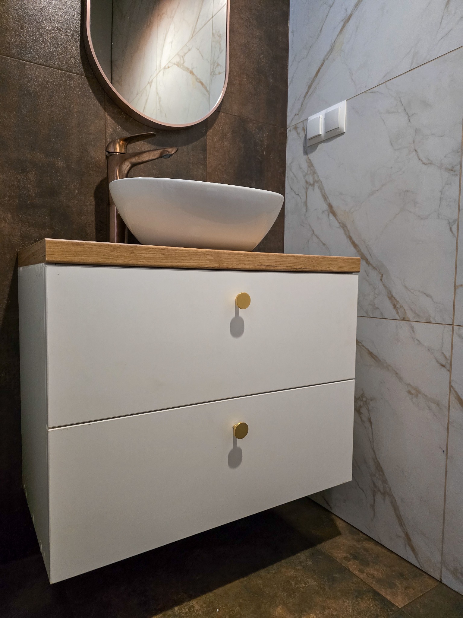 Biała szafka łazienkowa z drewnianym blatem, owalna umywalka i miedziana armatura na tle brązowych i marmurowych płytek. Złote gałki.