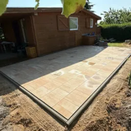 Ułożona, prostokątna nawierzchnia z jasnych płyt brukowych z ramą, otoczona piaskiem. W tle drewniany domek z oknami i zielona roślinność.