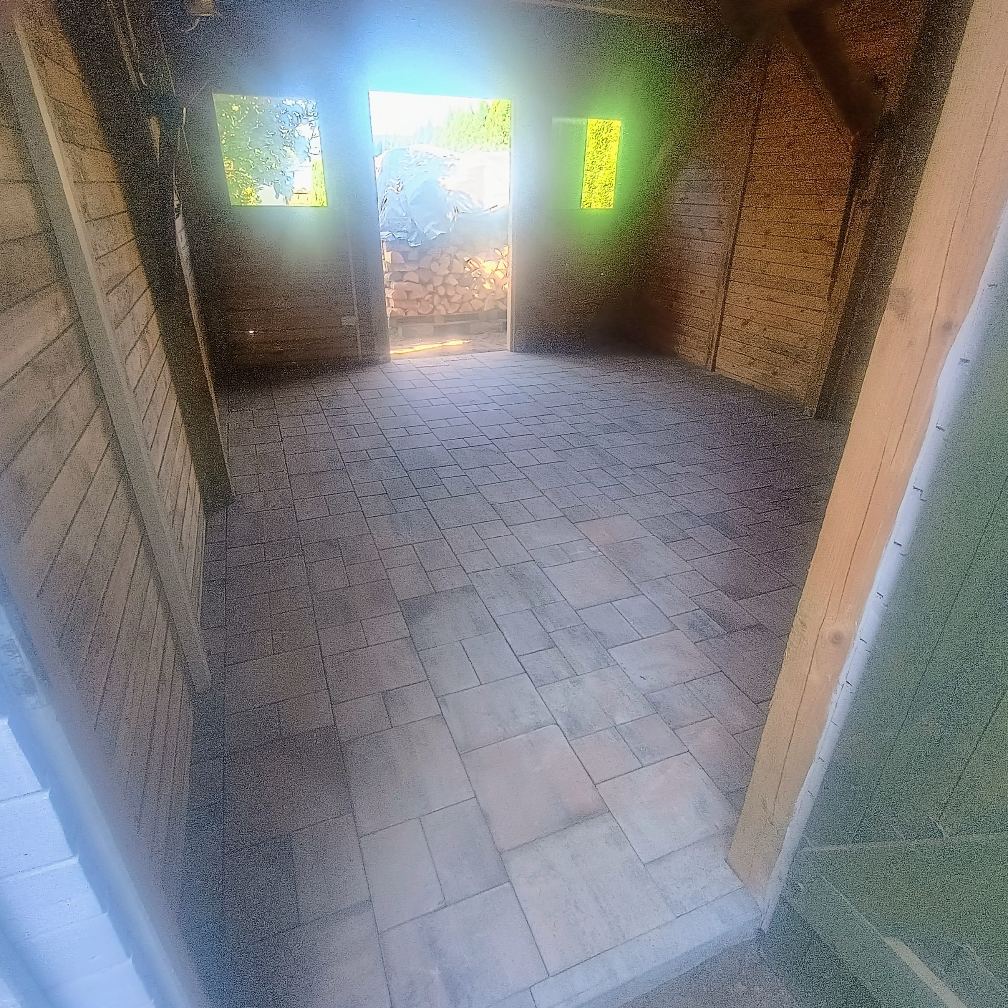 Ułożona kostka brukowa wewnątrz drewnianej konstrukcji. Widoczne regularne wzory kostki i drewniane ściany. W tle stos drewna.