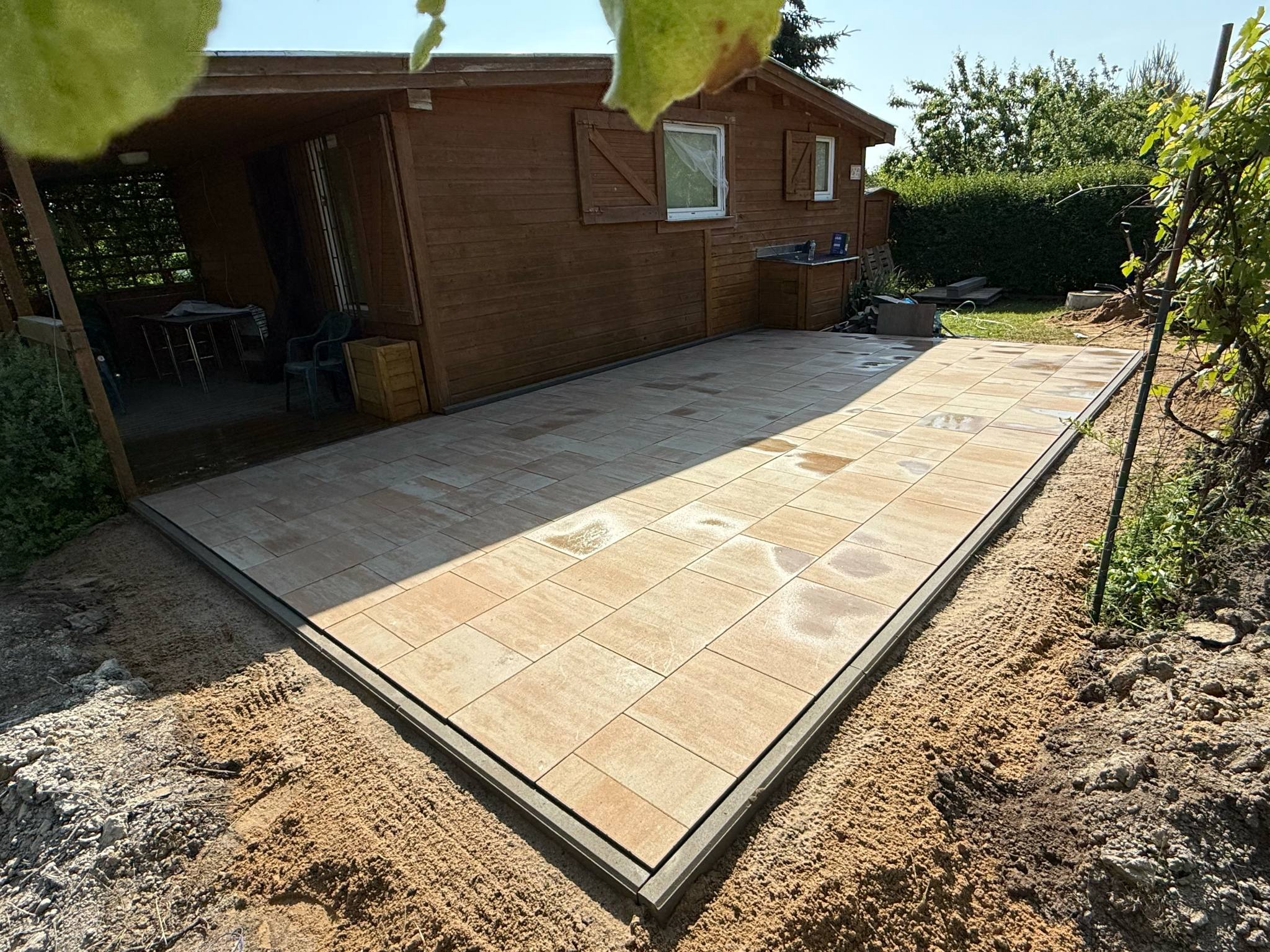 Ułożona, prostokątna nawierzchnia z jasnych płyt brukowych z ramą, otoczona piaskiem. W tle drewniany domek z oknami i zielona roślinność.