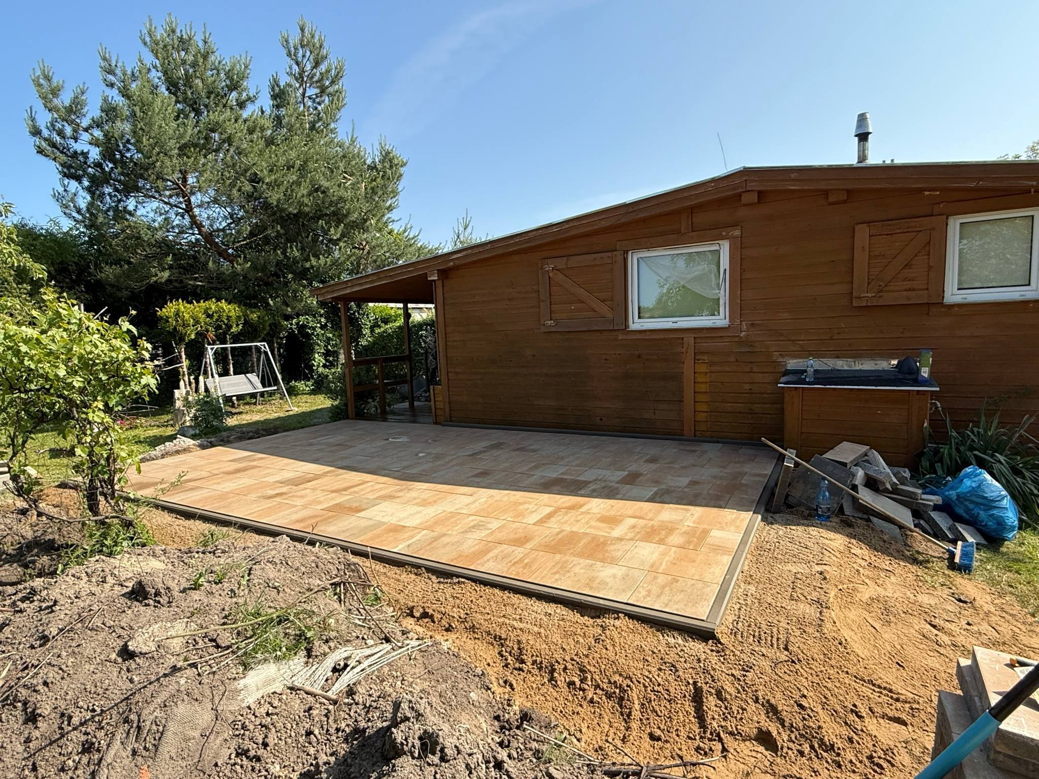 Ułożona kostka brukowa na tarasie przy drewnianym domku letniskowym. Widoczne narzędzia i materiały budowlane. Jasne, słoneczne oświetlenie.