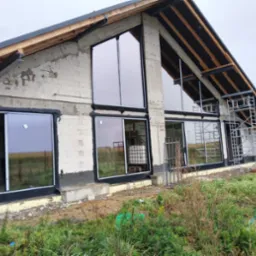 Surowy budynek z dużymi oknami z czarnymi ramami w trakcie montażu. Widoczne rusztowania i elementy konstrukcyjne. Tło stanowi pole i pochmurne niebo.