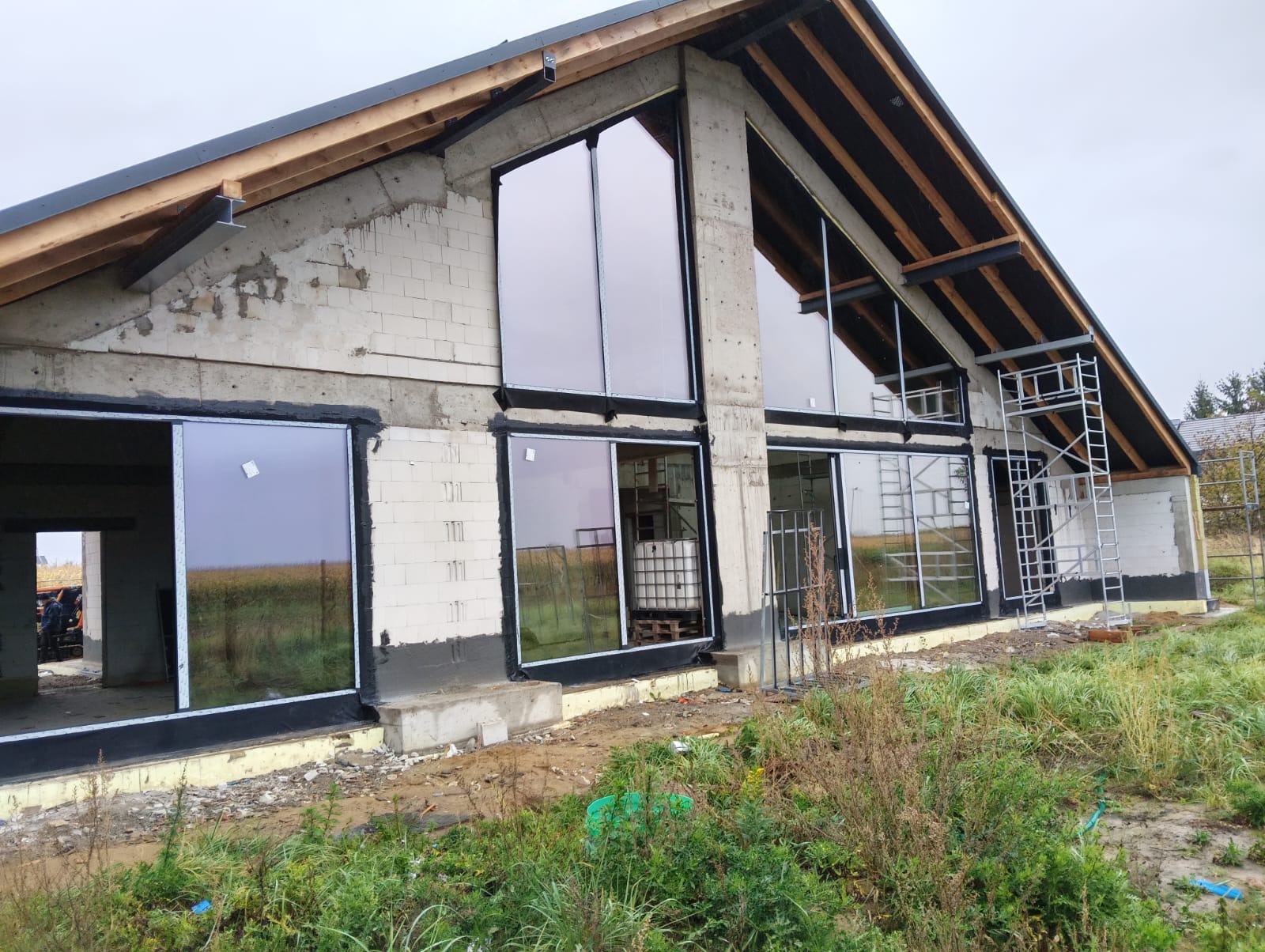 Surowy budynek z dużymi oknami z czarnymi ramami w trakcie montażu. Widoczne rusztowania i elementy konstrukcyjne. Tło stanowi pole i pochmurne niebo.