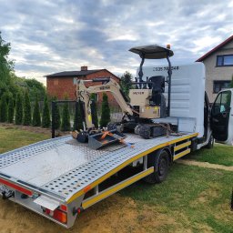 DIDA 2 SPÓŁKA Z OGRANICZONĄ ODPOWIEDZIALNOŚCIĄ - Mini koparka Meyer EX10 zabezpieczona pasami na platformie transportowej białego busa. Widok na dom i drzewa w tle, pochmurne niebo.