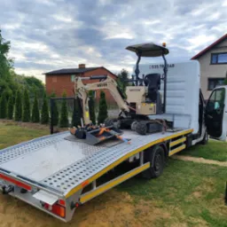 Mini koparka Meyer EX10 zabezpieczona pasami na platformie transportowej białego busa. Widok na dom i drzewa w tle, pochmurne niebo.