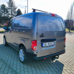 Szary Nissan NV200 z bagażnikiem dachowym, widok z tyłu na brukowanym parkingu w słoneczny dzień. Na klapie naklejka motocyklisty i tablica rejestracyjna.