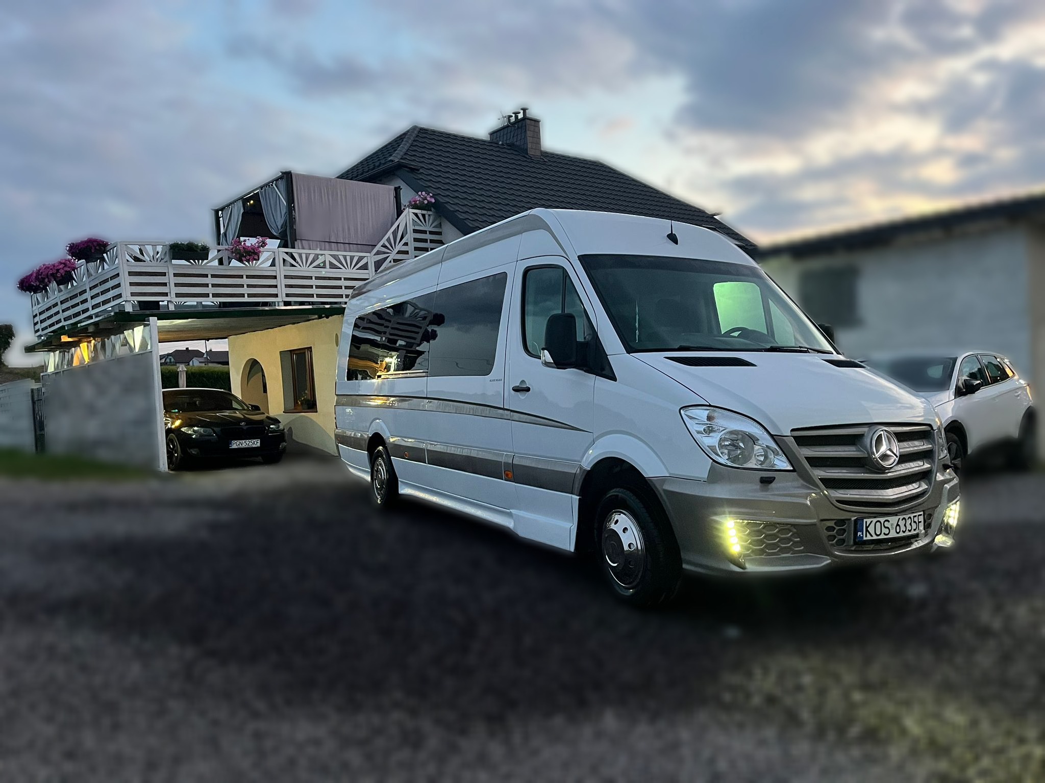 Biały Mercedes Sprinter z włączonymi światłami do jazdy dziennej, zaparkowany na tle domu z balkonem i dwoma samochodami osobowymi.
