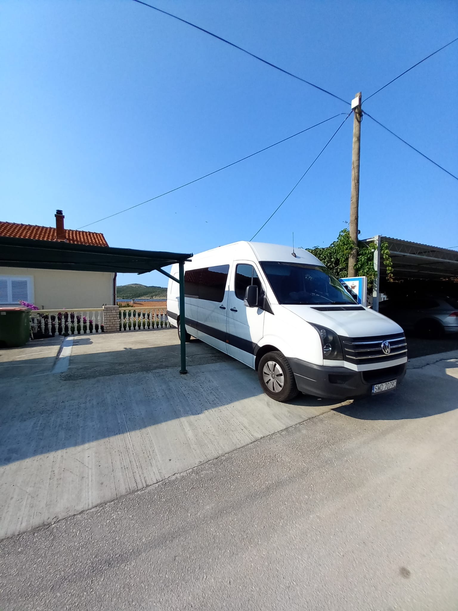 Biały bus parkuje w cieniu w słoneczny dzień. Widoczny numer rejestracyjny SWD 7079C, część budynku z czerwonym dachem i błękitne niebo w tle.