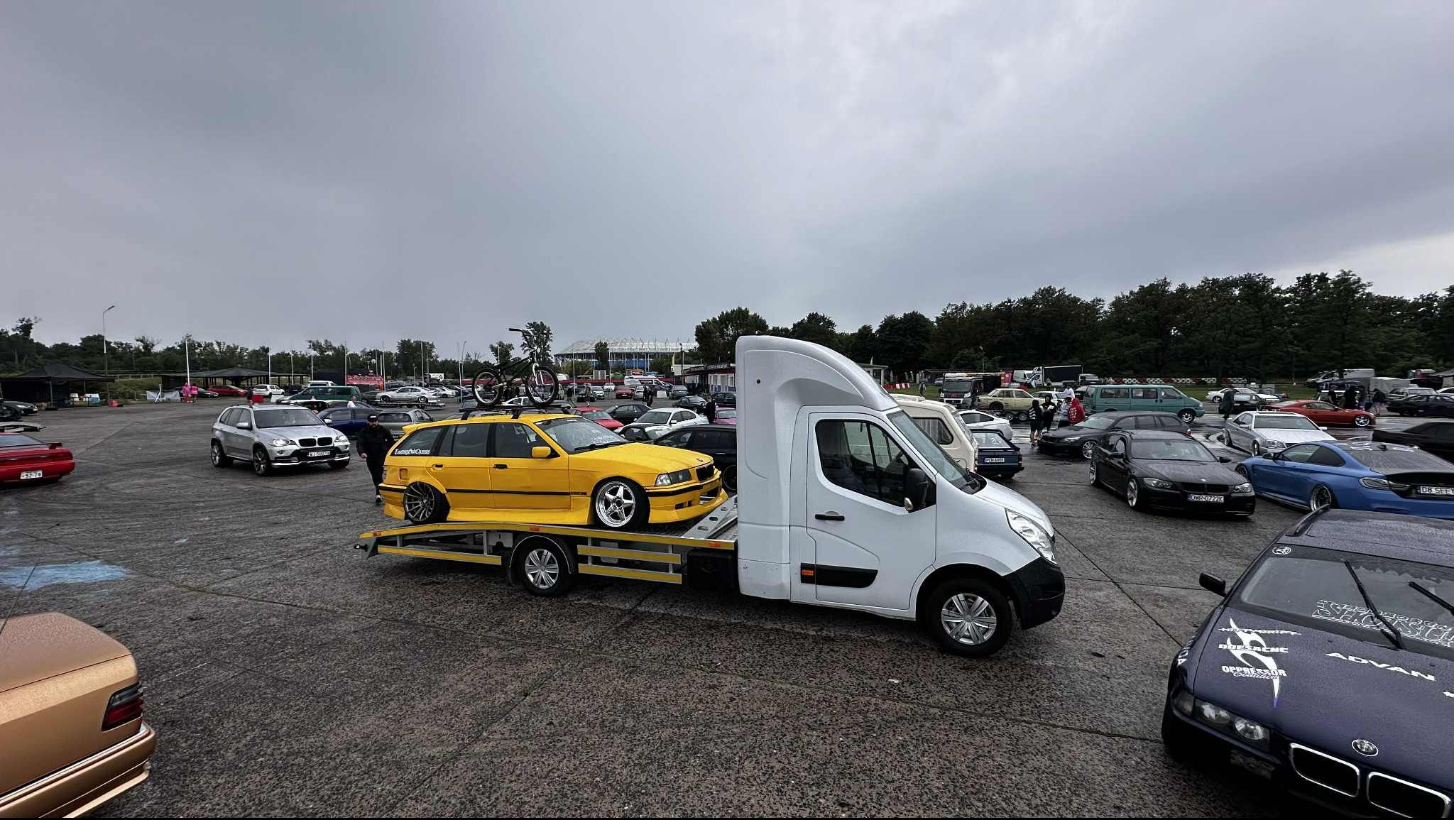 Żółte BMW na lawecie Renault Master na tle parkingu pełnego aut podczas zlotu miłośników motoryzacji. Na dachu BMW zamocowany rower. Pochmurne niebo.