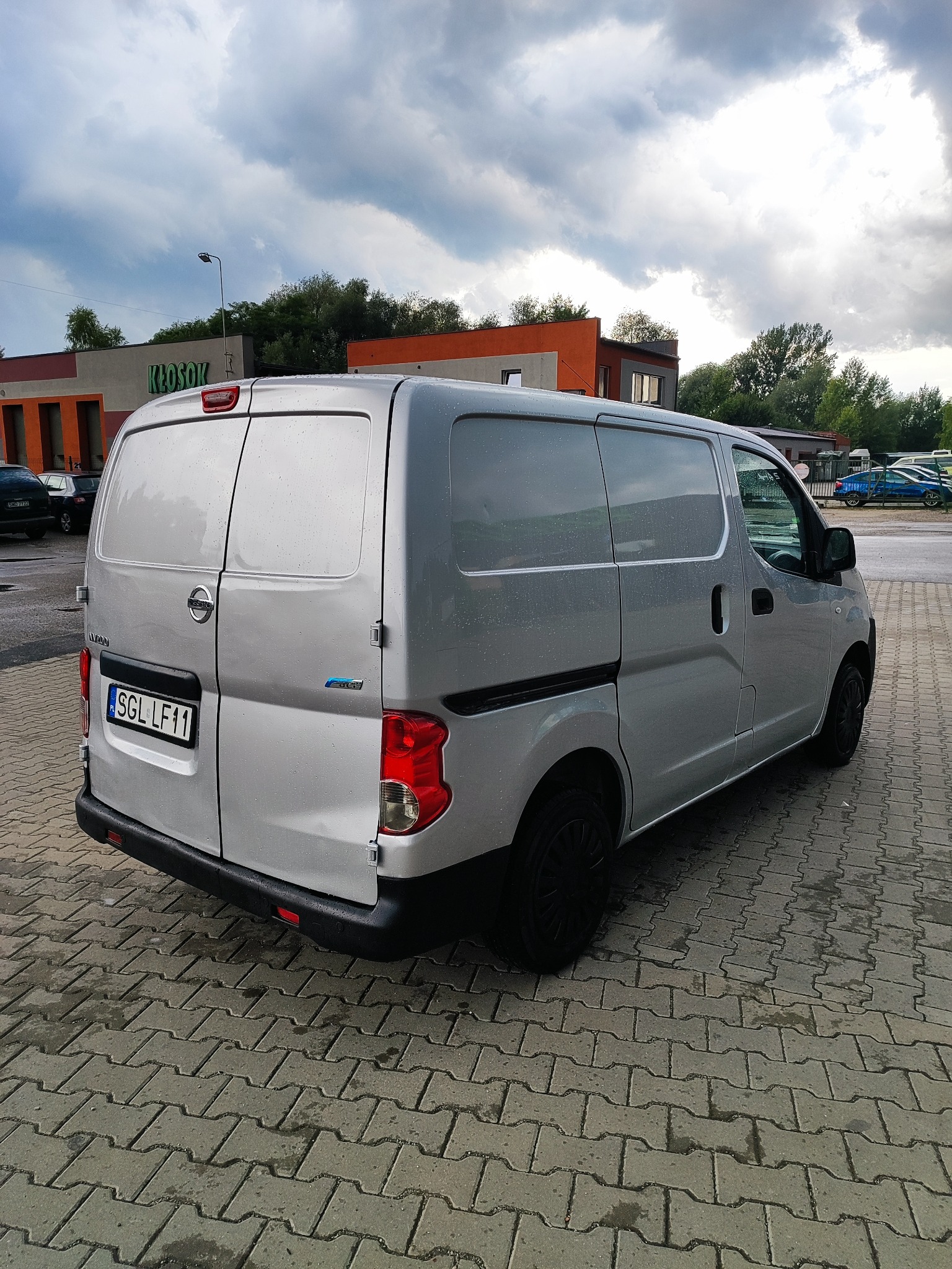 Srebrny, dostawczy Nissan NV200 z czarnymi felgami, zaparkowany na kostce brukowej. Widok z tyłu, na tle budynku i pochmurnego nieba.