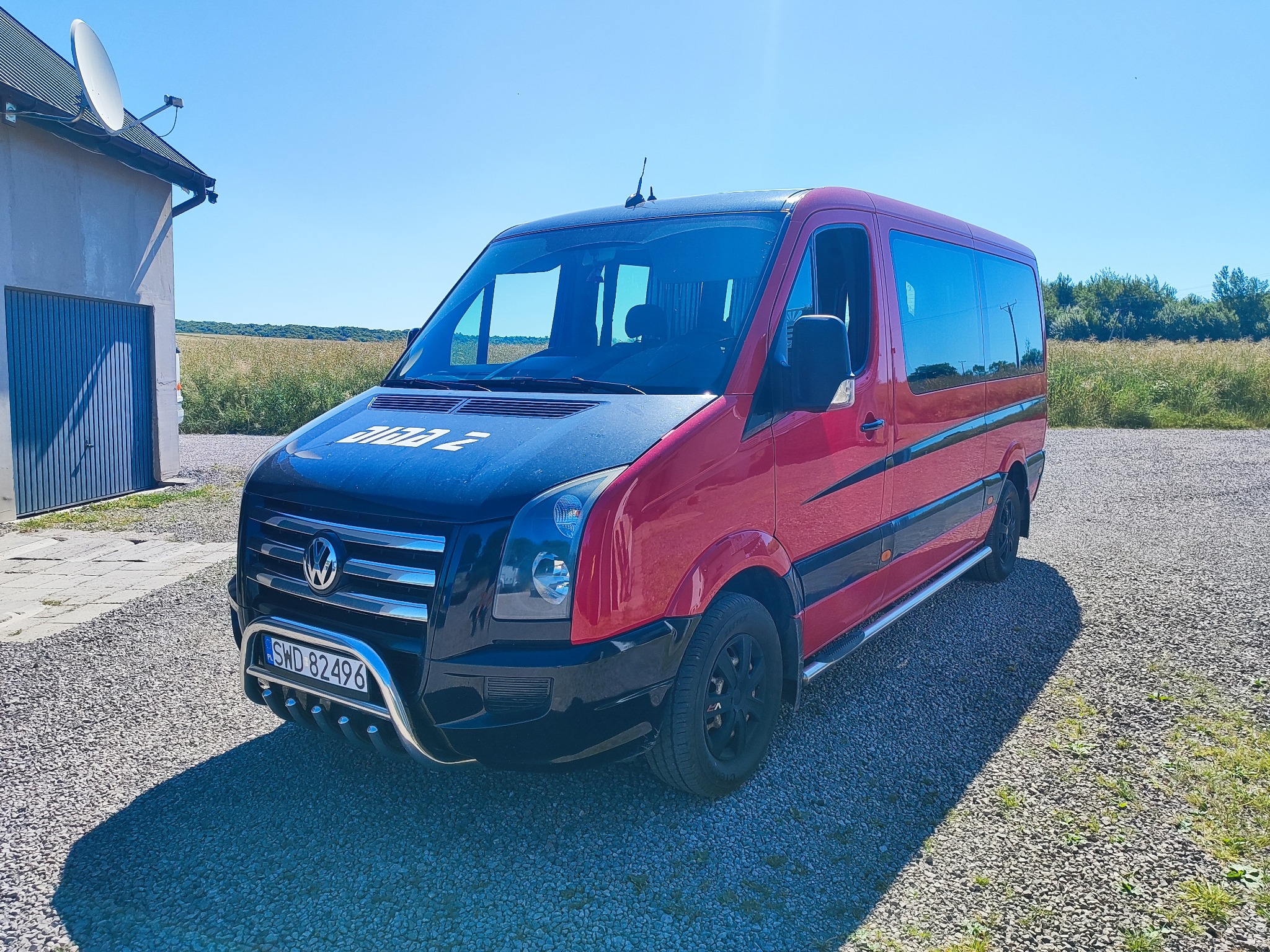 Czerwono-czarny bus VW Crafter z napisem 'Dizzy 2' na masce, zaparkowany na żwirowym podłożu, z budynkiem i polem w tle, w słoneczny dzień.