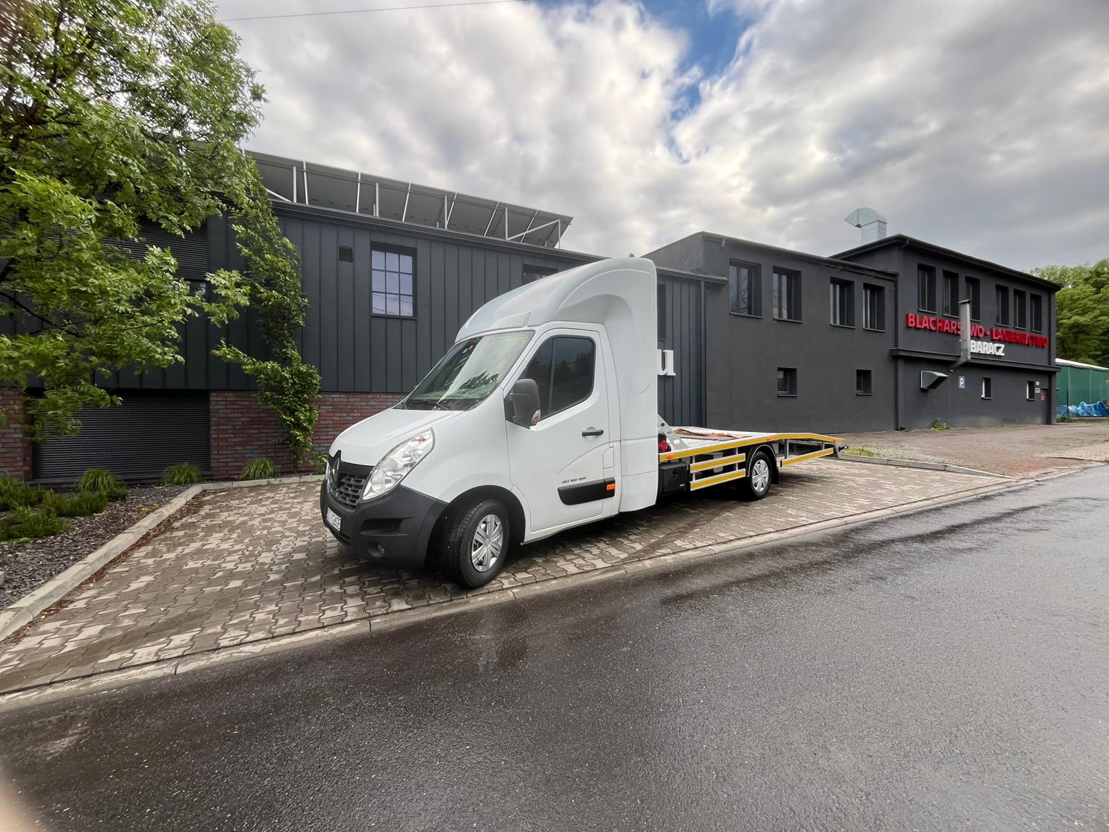 Biała laweta Renault Master z żółtymi akcentami na platformie, zaparkowana przed budynkiem warsztatu blacharsko-lakierniczego w pochmurny dzień.