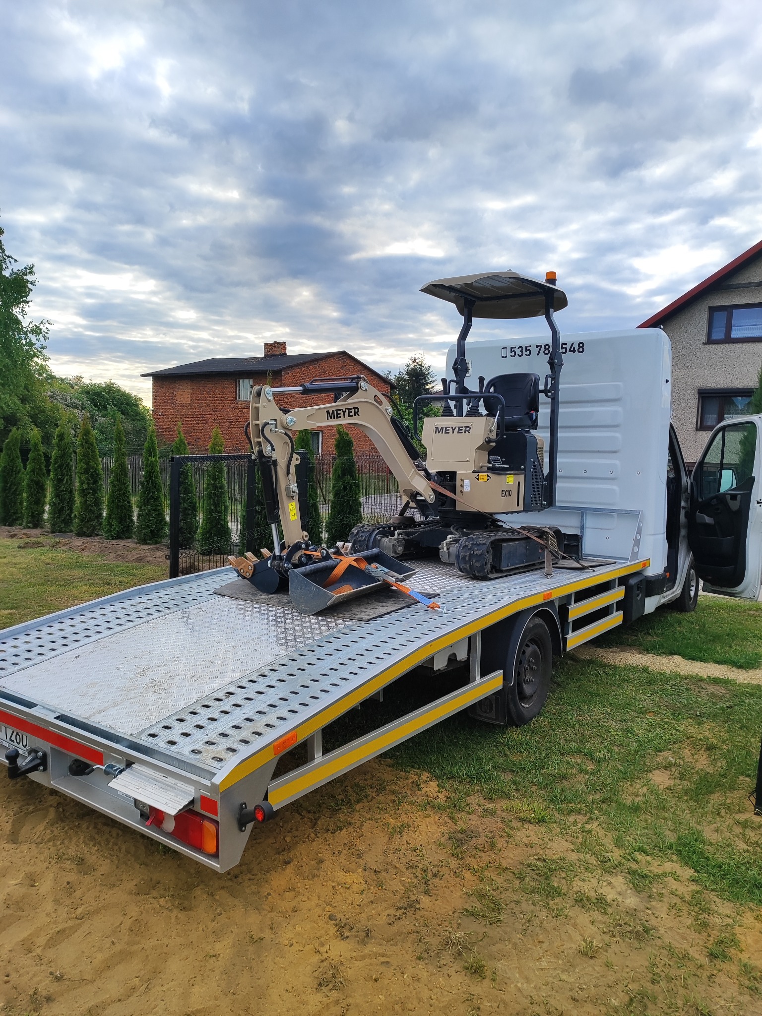 Mini koparka Meyer EX10 zabezpieczona pasami na platformie transportowej białego busa. Widok na dom i drzewa w tle, pochmurne niebo.