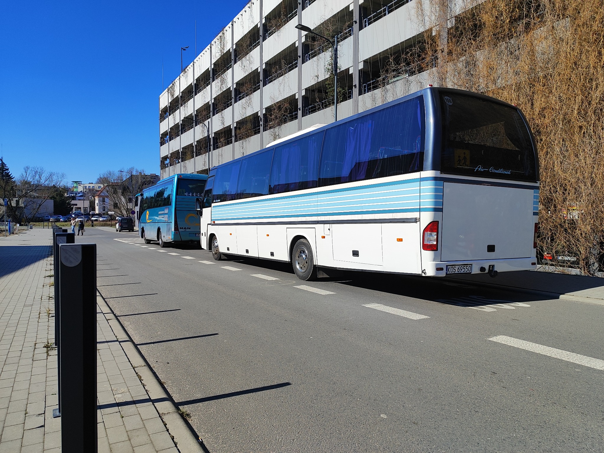 Dwa autokary turystyczne zaparkowane na ulicy w słoneczny dzień, z widocznym budynkiem parkingu w tle. Biało-niebieski autokar z tablicą rejestracyjną na pierwszym planie.