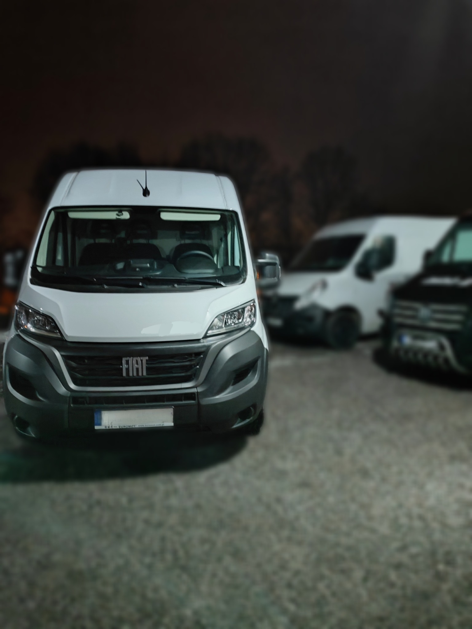 Biały Fiat Ducato na parkingu nocą, z widocznym logo i reflektorami, w tle inne busy. Ujęcie z przodu, lekko z dołu, podkreślające wielkość pojazdu.