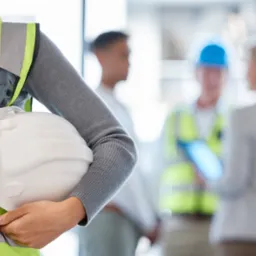 Osoba w kamizelce odblaskowej trzyma kask na budowie, w tle rozmawiający pracownicy. Bezpieczeństwo pracy, szkolenia BHP na budowie.
