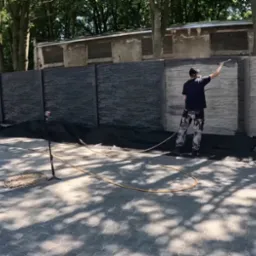 Malowanie betonowego ogrodzenia natryskowo. Część ogrodzenia pomalowana na ciemny kolor, druga część w odcieniach szarości. Prace w toku, widoczny pracownik i sprzęt.