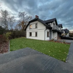 Nowoczesny dom z zadbanym trawnikiem i kostką brukową. Widok na elewację, regularnie skoszony trawnik z opadłymi liśćmi i fragment podjazdu z ciemnej kostki.