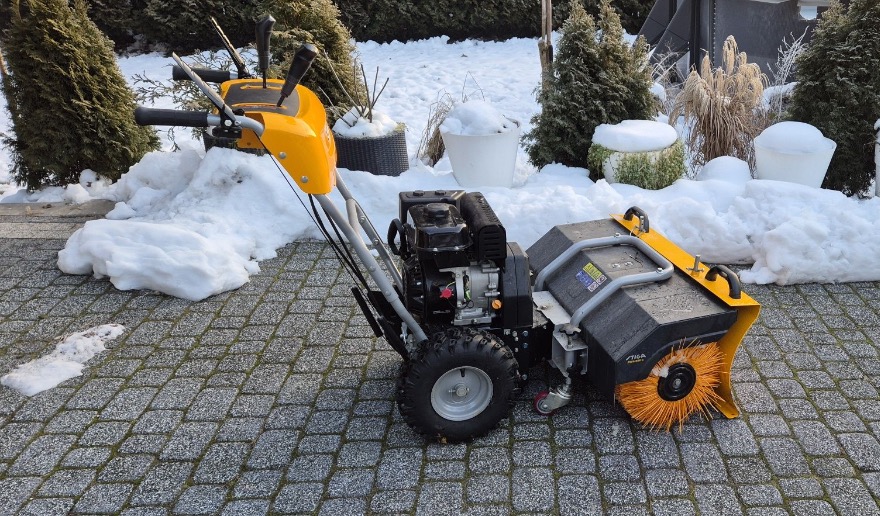 Żółto-czarna zamiatarka spalinowa Stiga na kostce brukowej, w tle zaśnieżone krzewy i donice. Sprzęt do odśnieżania dróg i placów.