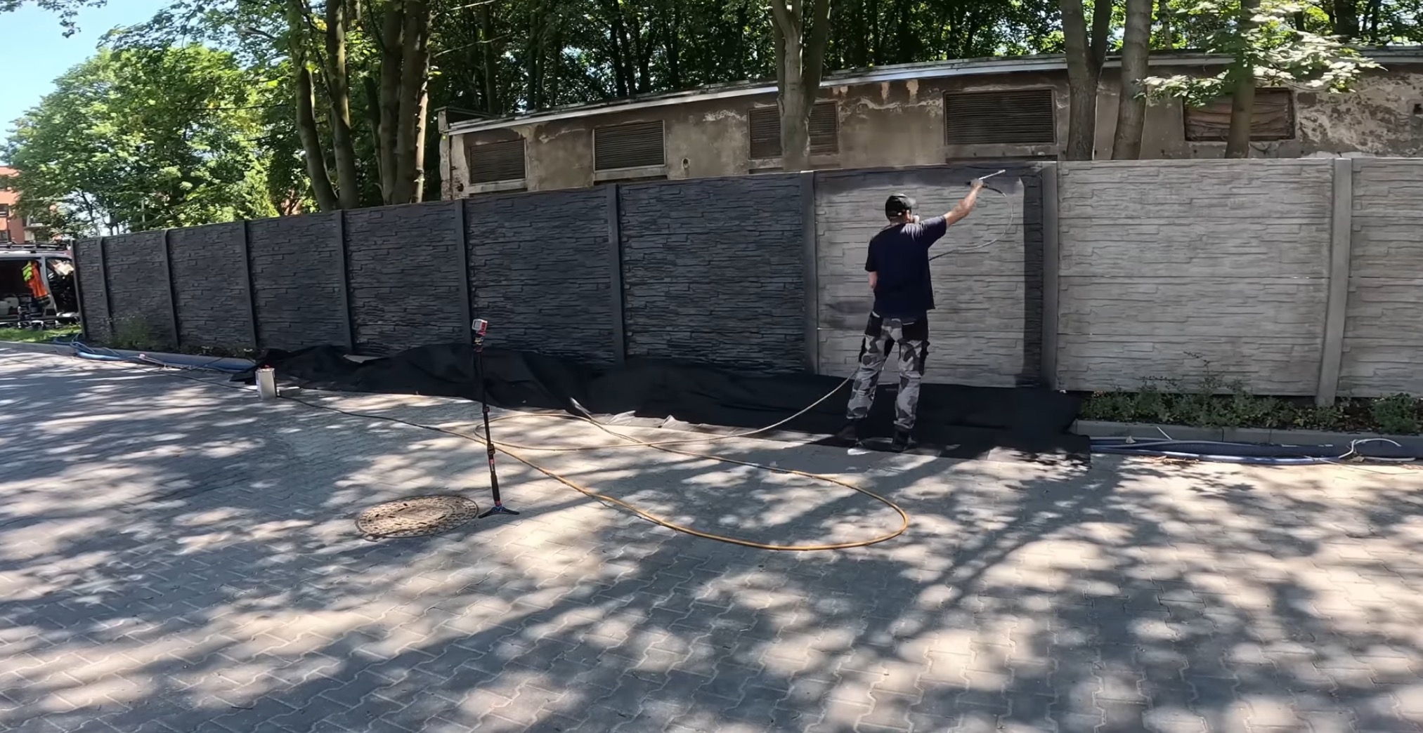 Malowanie betonowego ogrodzenia natryskowo. Część ogrodzenia pomalowana na ciemny kolor, druga część w odcieniach szarości. Prace w toku, widoczny pracownik i sprzęt.