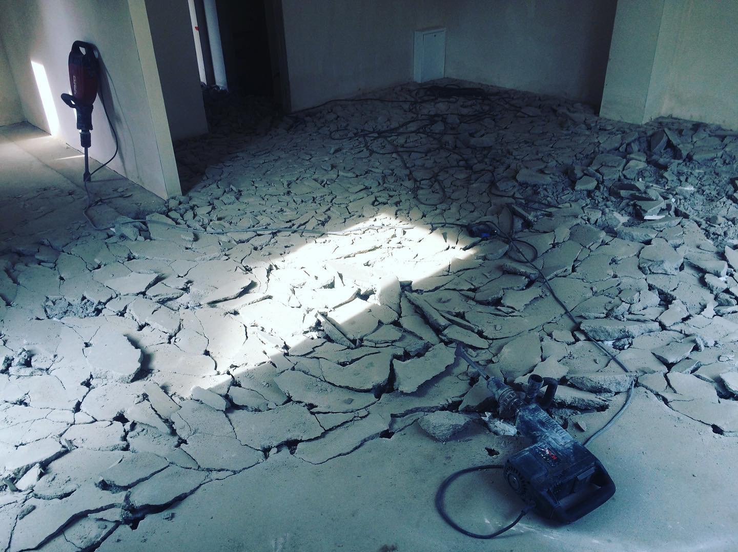 Demontaż posadzki w pomieszczeniu z użyciem młota udarowego. Widoczne fragmenty gruzu i narzędzia pracy. Jasne światło pada na podłogę.