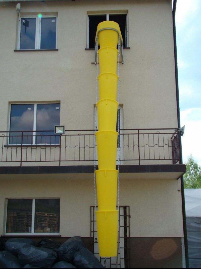 Żółty zsyp budowlany przymocowany do okna budynku, służący do usuwania gruzu. Widoczne czarne worki na dole. Ujęcie z dołu, na tle jasnej elewacji.