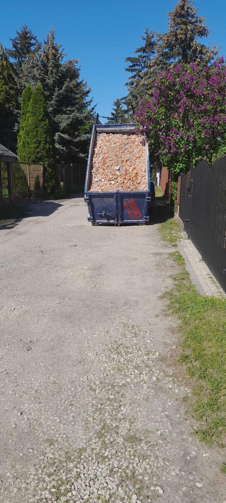 Kontener wypełniony gruzem na żwirowej drodze, otoczony zielenią drzew i krzewów. Widoczny fragment drewnianego i metalowego ogrodzenia.