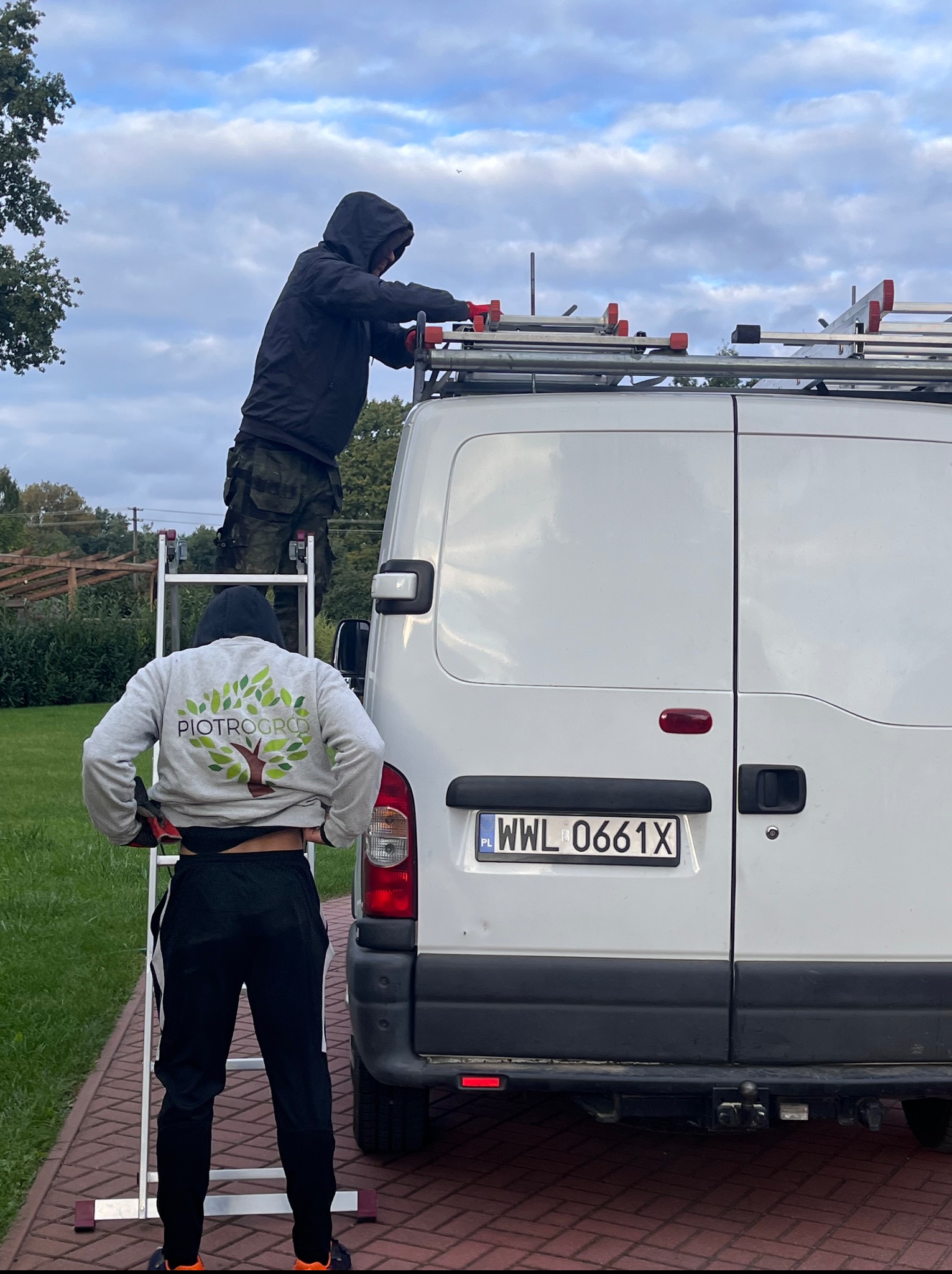 Pracownik na drabinie mocuje sprzęt na dachu białego busa z logo 'PIOTROGRÓD', drugi pracownik asekuruje drabinę. Usługi ogrodnicze.
