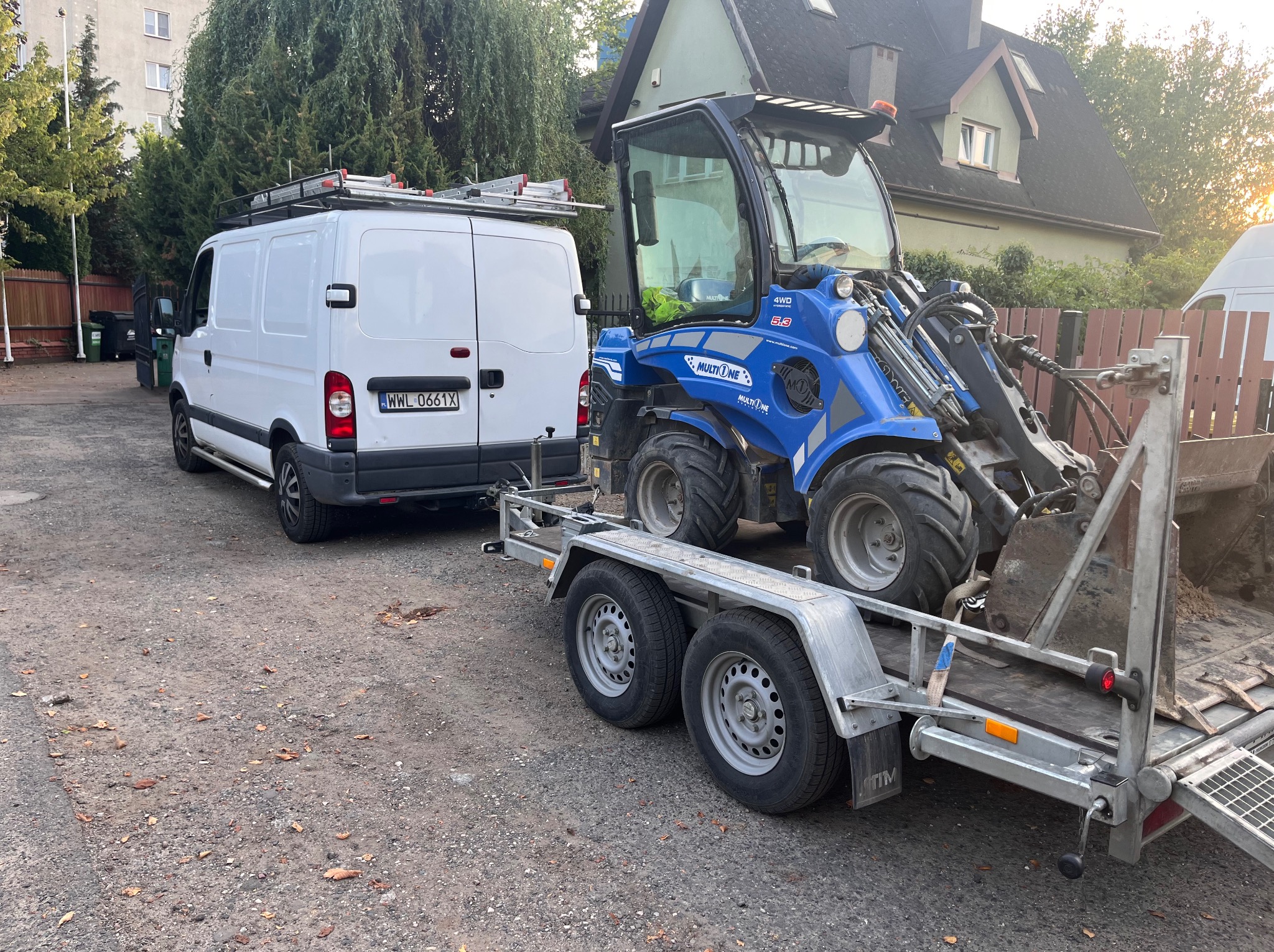 Biały van z drabinką na dachu i niebieska miniładowarka Multione na przyczepie, gotowe do pracy w ogrodzie. Dom w tle.