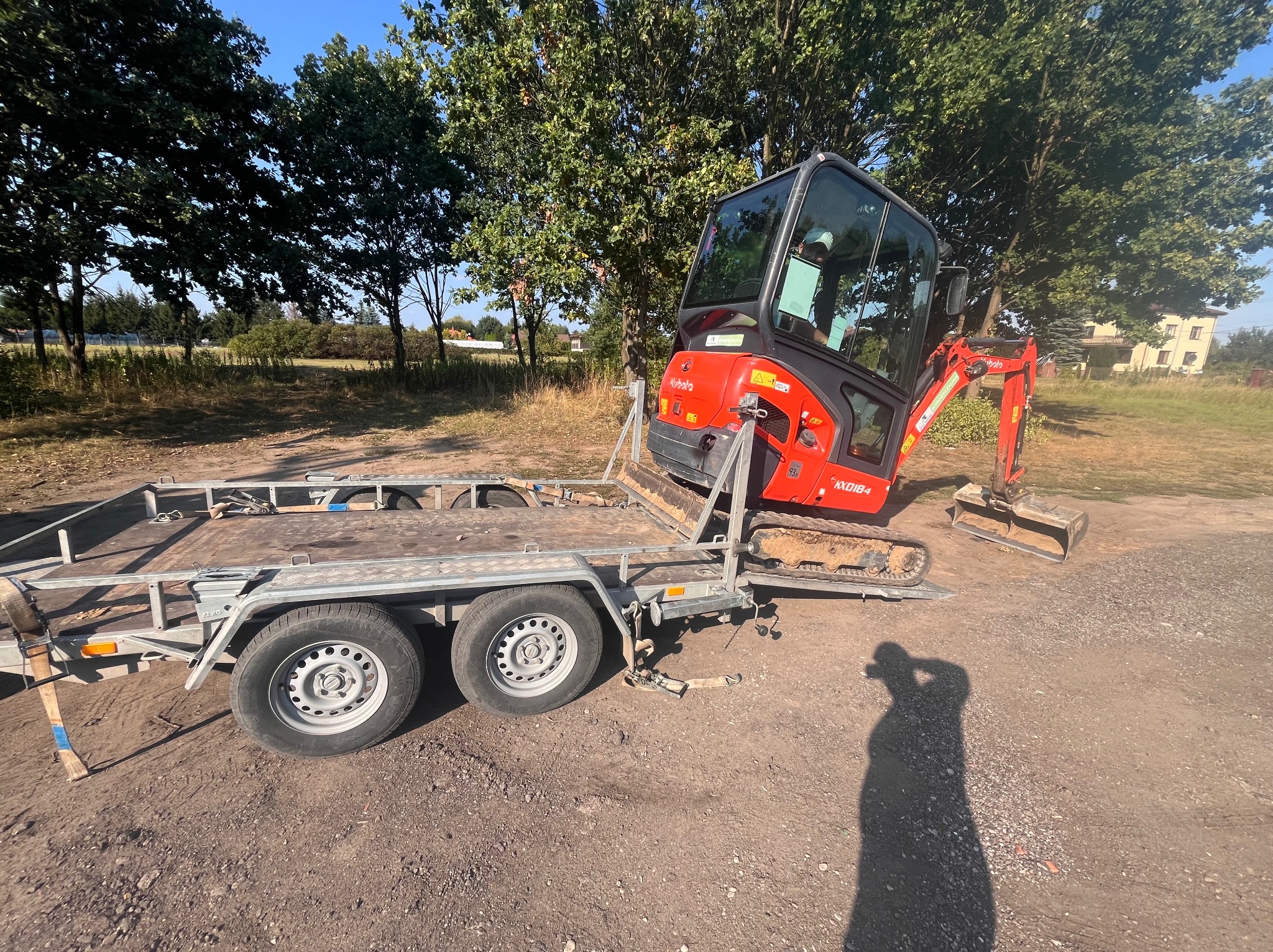 Czerwona mini koparka Kubota KX018-4 na przyczepie, gotowa do transportu, z widocznym otoczeniem zieleni i budynków. Słoneczny dzień.