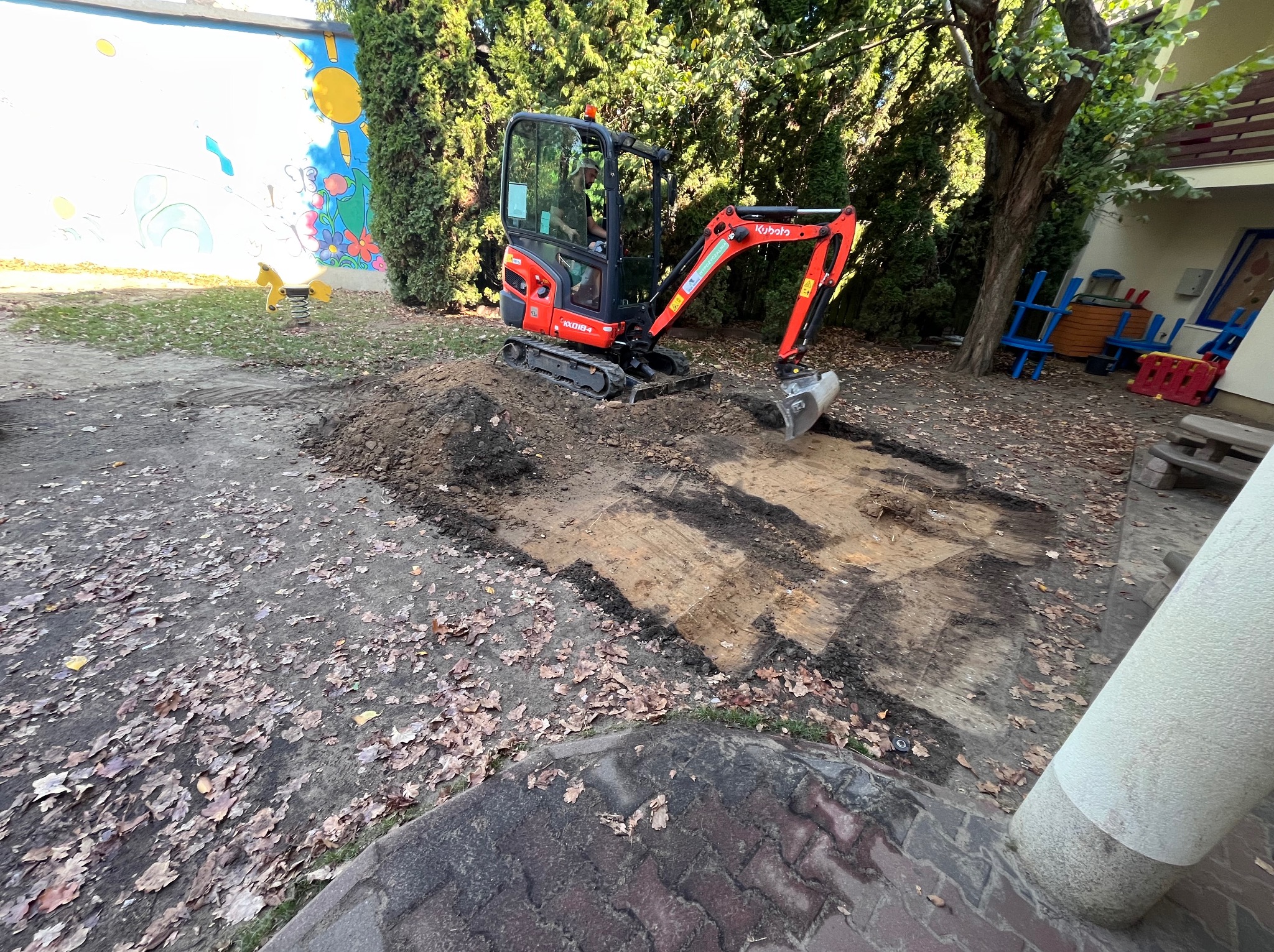 Czerwona mini koparka Kubota na placu budowy, wykopana ziemia, w tle budynek z kolorowym muralem i drzewa. Prace ziemne w toku.