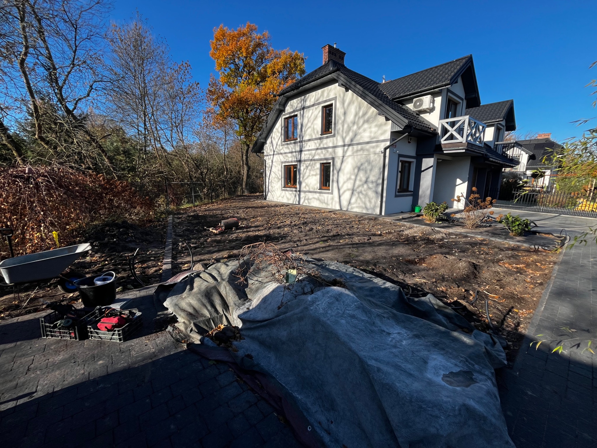 Przygotowany teren pod nasadzenia przed domem jednorodzinnym. Widoczne narzędzia ogrodnicze i plandeka. Jesienne słońce oświetla dom i drzewo z żółtymi liśćmi.