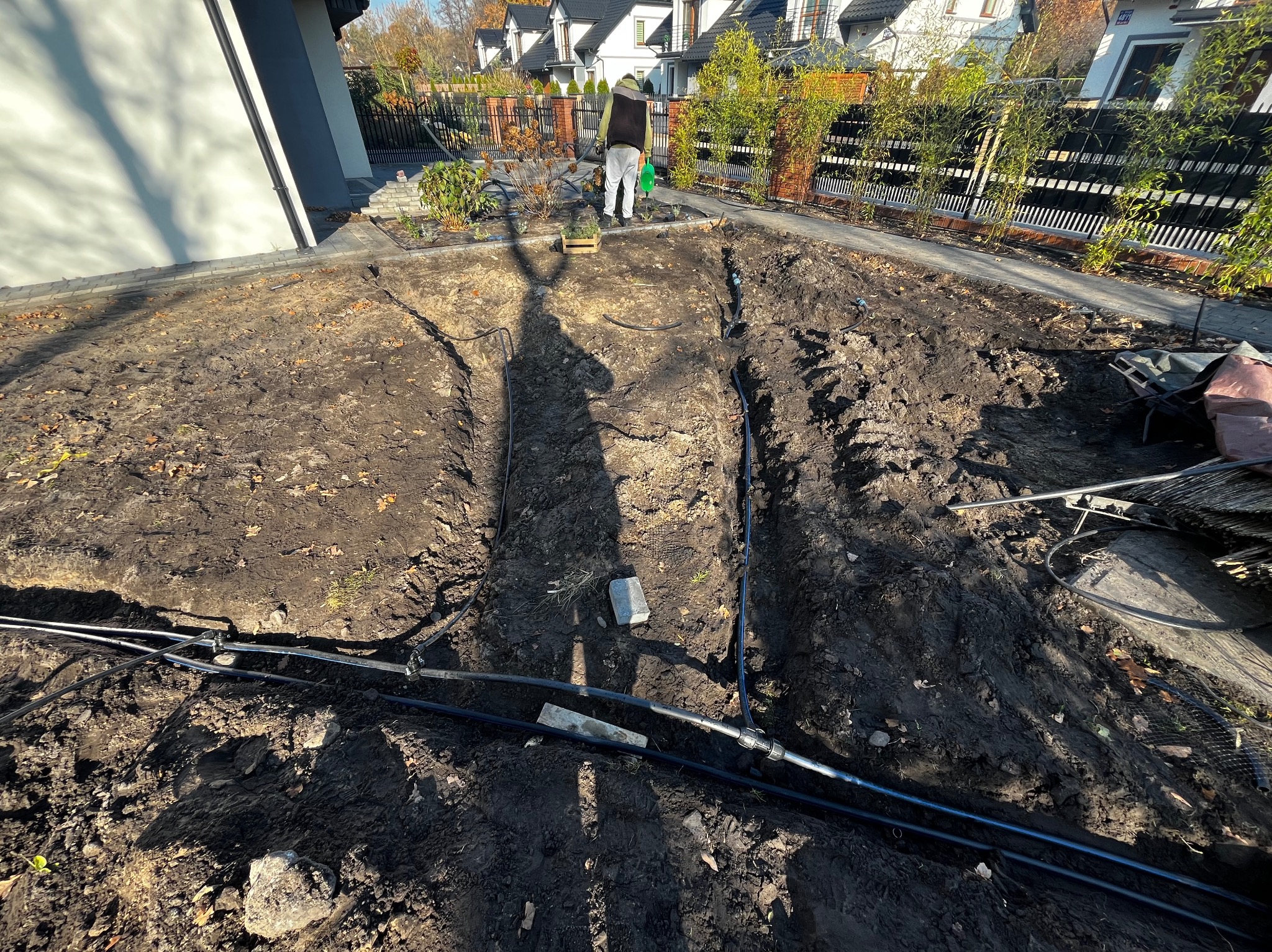 Instalacja systemu nawadniania w ogrodzie: czarne rury w wykopanych rowach, ogrodnik z konewką w tle. Widok z góry na przygotowany teren.