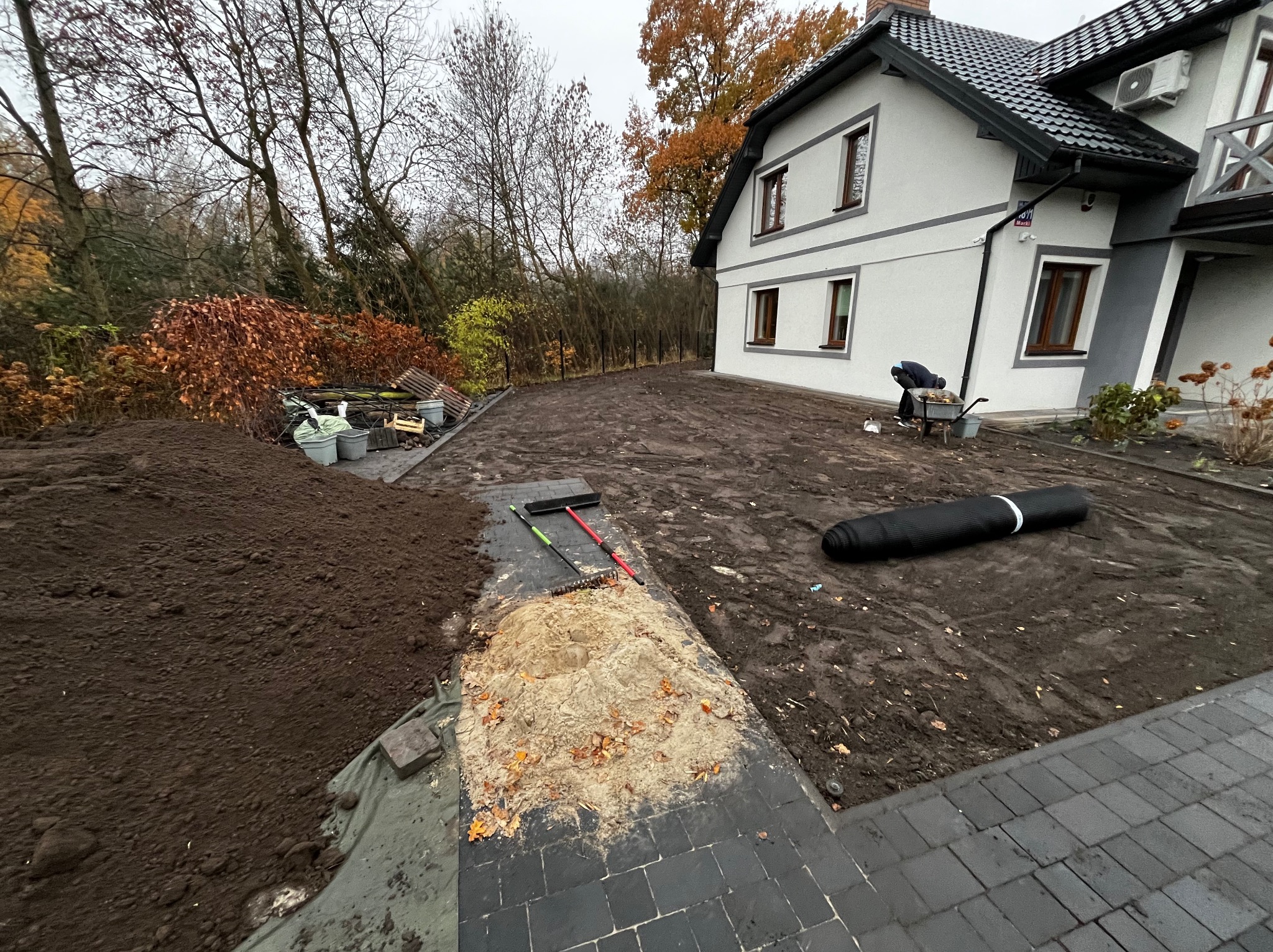 Przygotowanie terenu pod trawnik: wyrównana ziemia, hałdy ziemi, narzędzia ogrodnicze i siatka, dom w tle. Prace ziemne w ogrodzie przed założeniem trawnika.